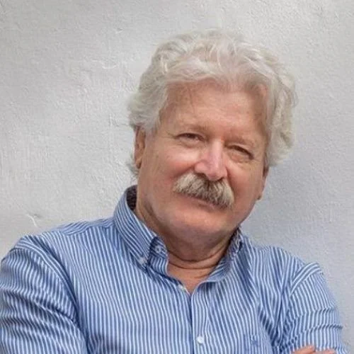 Hombre de cabello gris y bigote, con camisa a rayas azul, sonriendo y cruzando los brazos frente a una pared blanca.