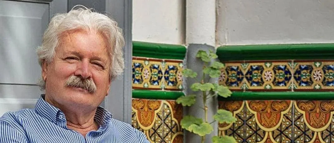 Hombre de cabello canoso y barba, con camiseta a rayas, sentado junto a un muro decorado con cerámica colorida y plantas.