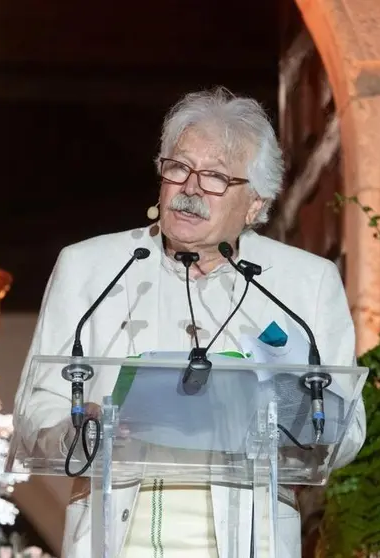 Un hombre mayor con gafas alzando la vista, vestido de blanco, parando en un podio con micrófonos en un entorno interior o en un escenario.