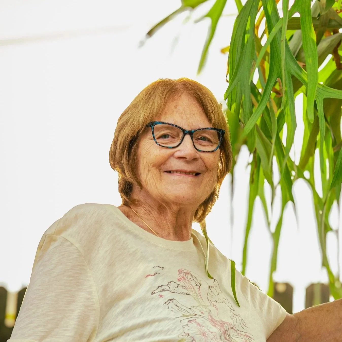 A smiling elderly woman with light brown hair, glasses, and fair skin, standing outdoors next to green foliage.