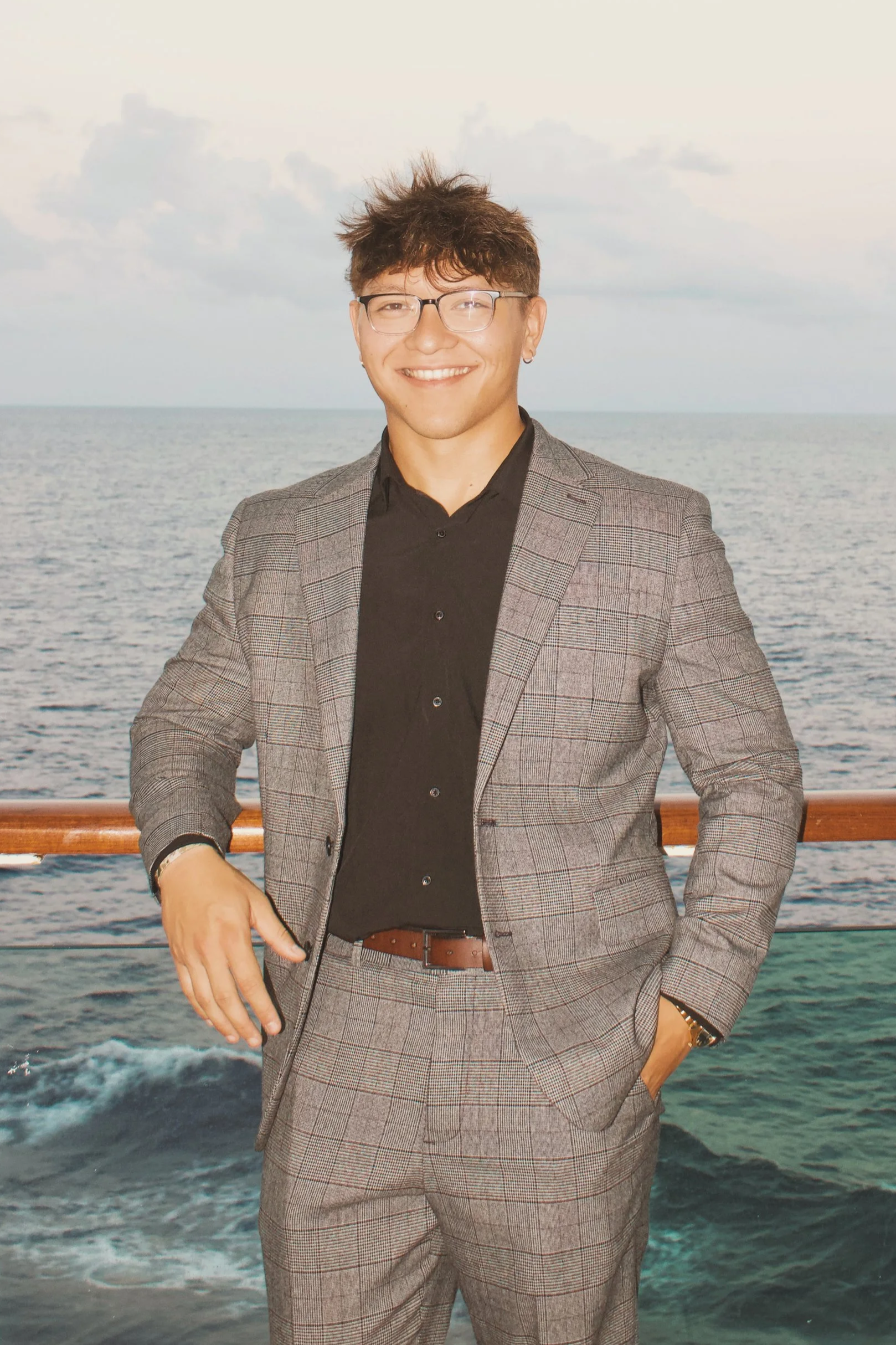 A man in a gray suit and black shirt stands on a boat or cruise ship deck, with the ocean in the background, smiling at the camera.