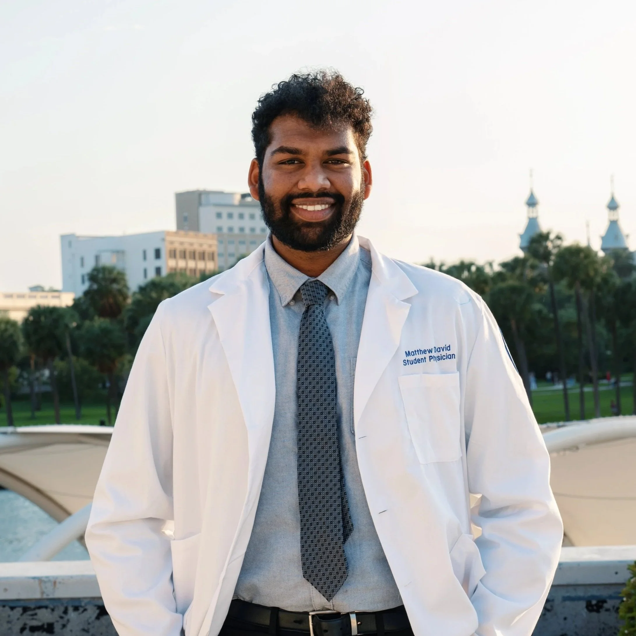 A man with dark curly hair and a beard, wearing a white lab coat with the name tag 'Matthew David, Student Physician', a light blue shirt, and a dark patterned tie, standing outdoors with a cityscape and two church steeples in the background during daytime.