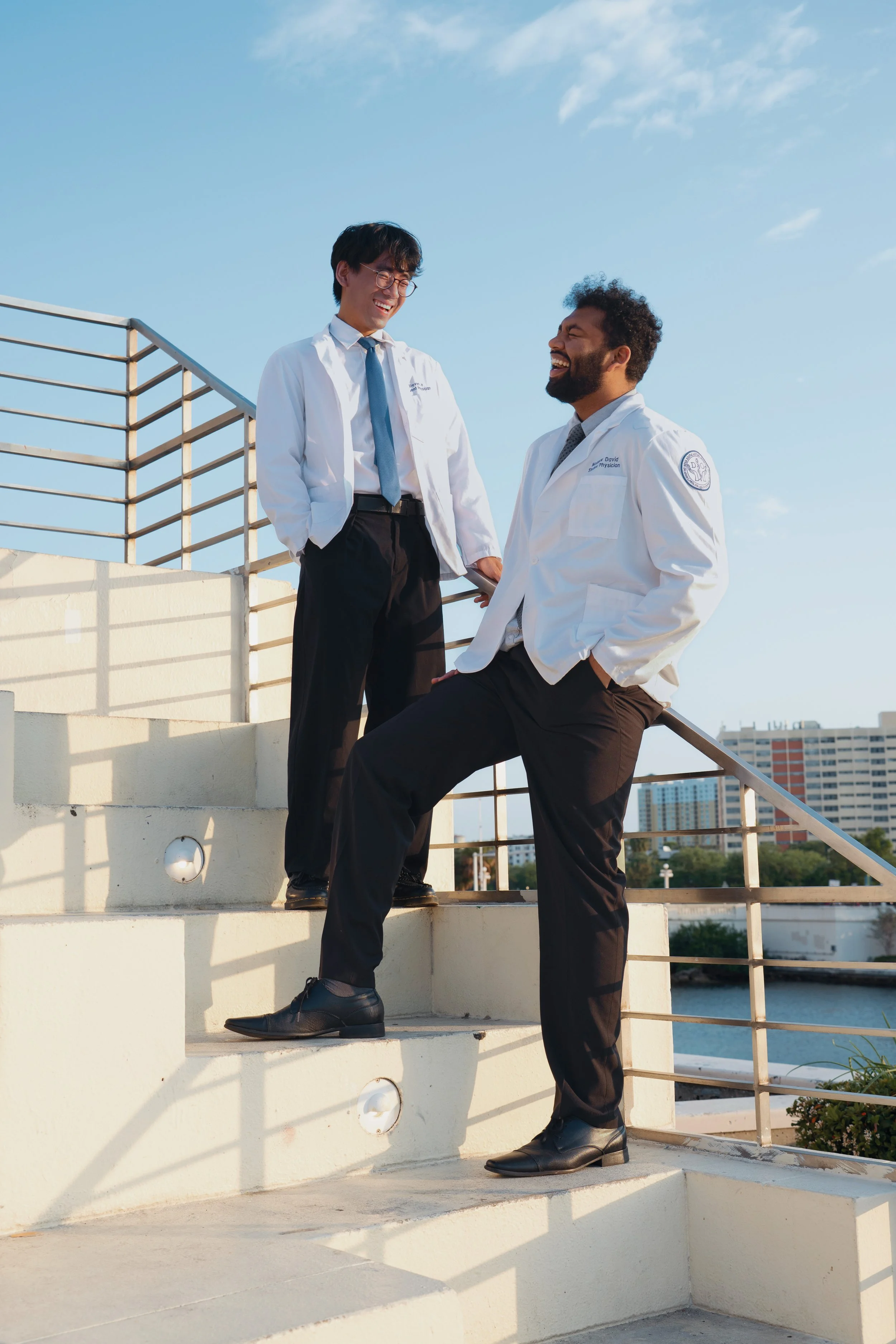 Two men in white lab coats standing on outdoor stairs, laughing and talking, with a city skyline and water in the background on a sunny day.