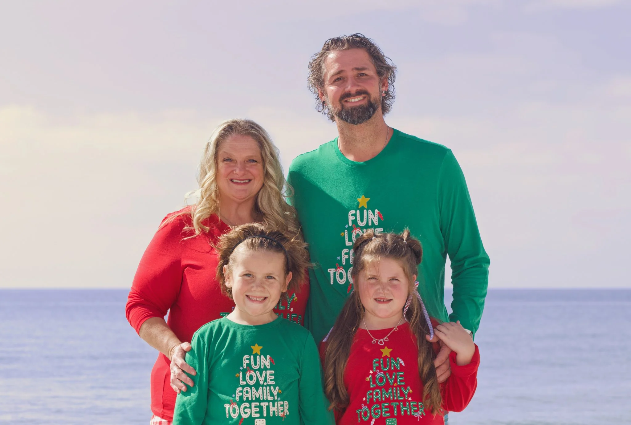 A smiling family of five outdoors by the ocean, dressed in red and green holiday sweaters with holiday messages, under a partly cloudy sky.