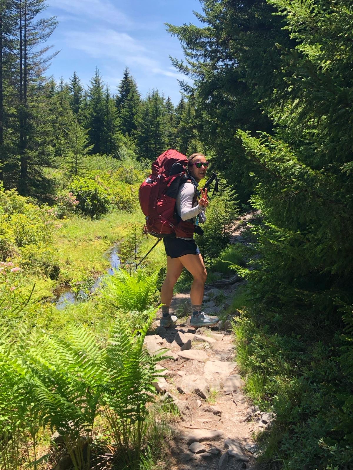 A woman hiking on a trail in a lush forest, carrying a large red backpack, wearing sunglasses, shorts, and hiking shoes, with tall green trees surrounding her.