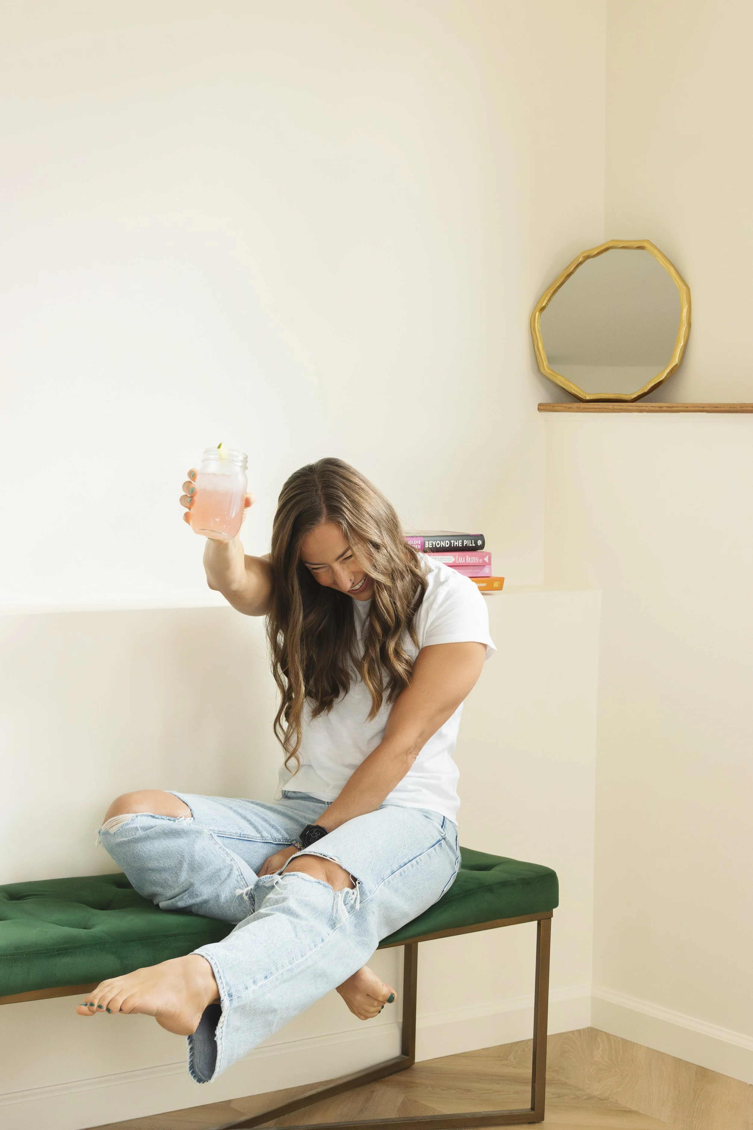 A young woman with long wavy brown hair, wearing a white t-shirt and ripped light blue jeans, sitting cross-legged on a cushioned bench, laughing and holding a mason jar with a pink drink.