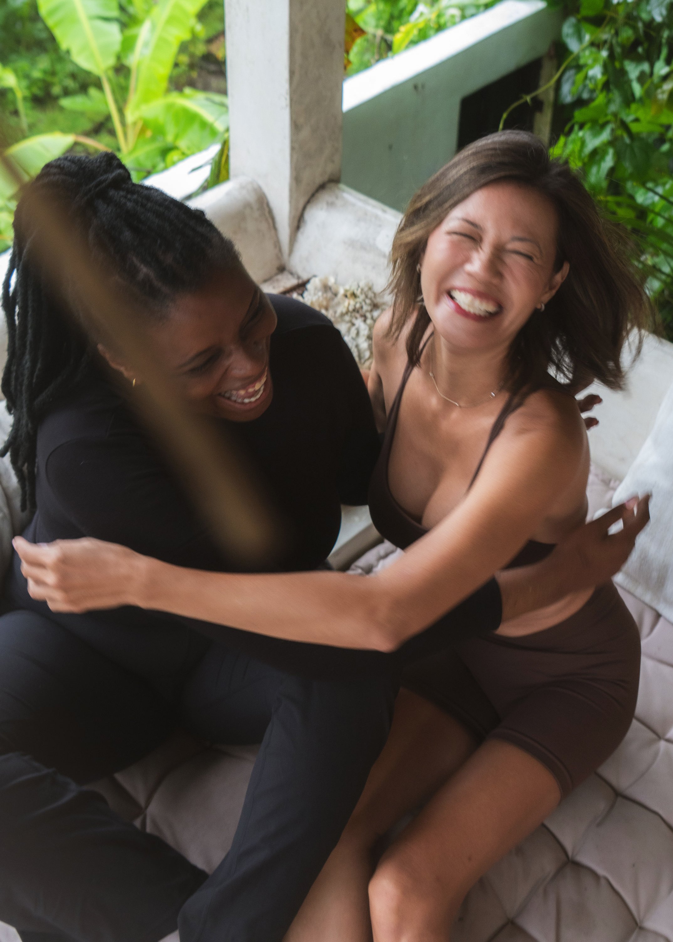 Two women sitting on a couch outdoors, hugging and laughing joyfully.