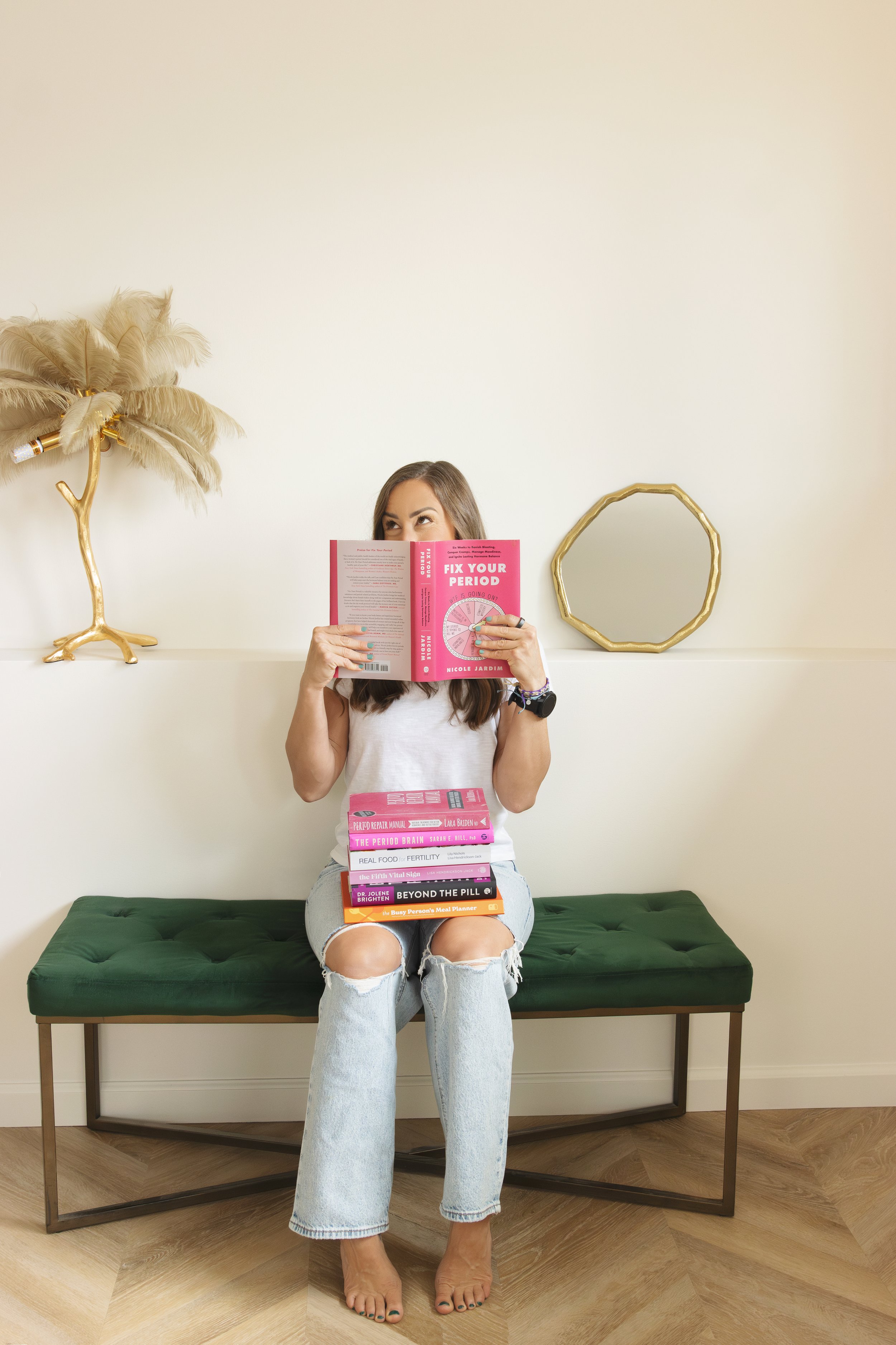 Woman sitting on a green cushioned bench, reading a book titled 'Fix Your Period'. She has a stack of similar books on her lap and wears ripped jeans and a sleeveless top. The room has decorative items on the wall, including a gold-colored branch with feathers and a geometric gold frame.