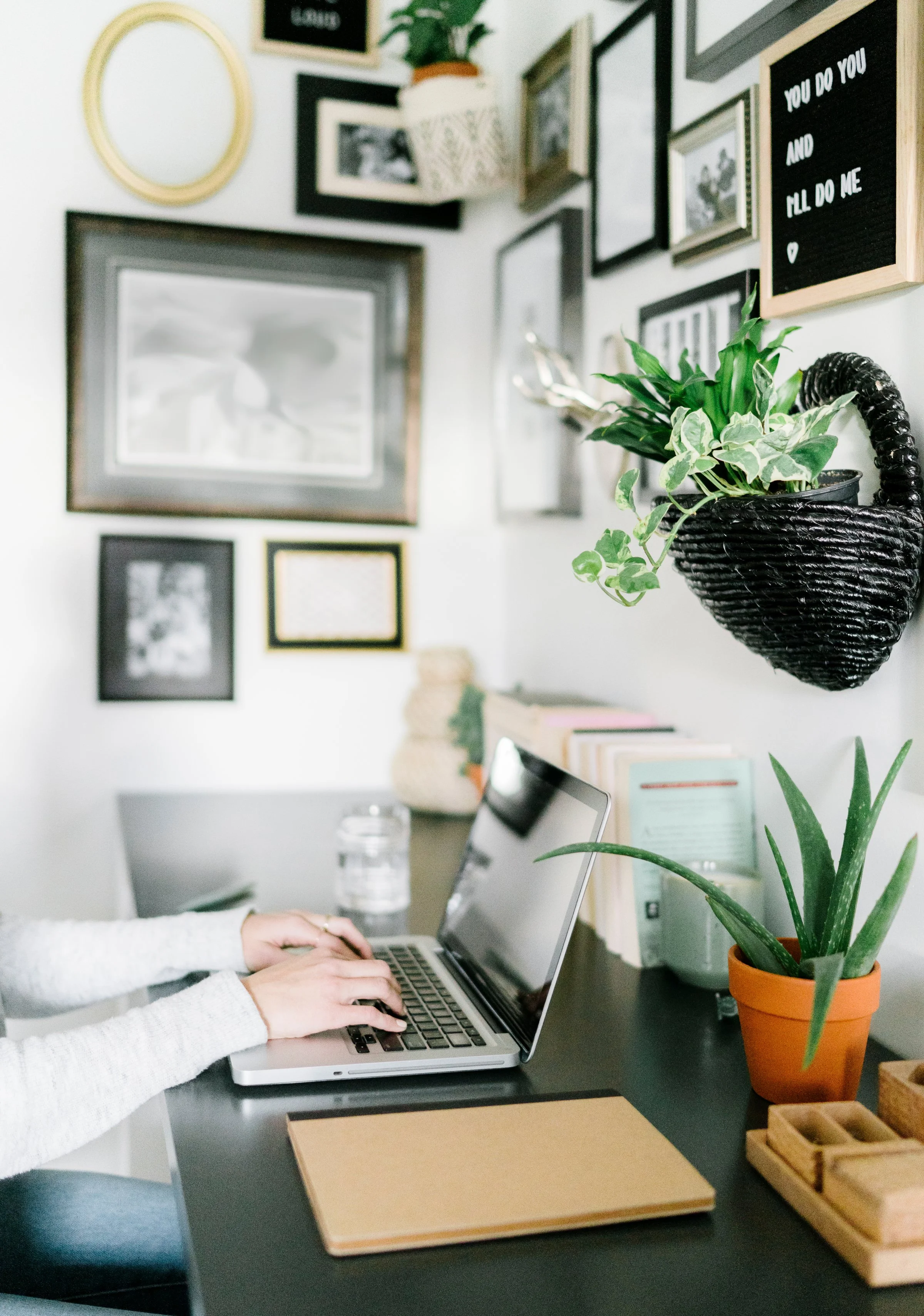 Person using laptop on a black desk with potted plants and picture frames on the wall.