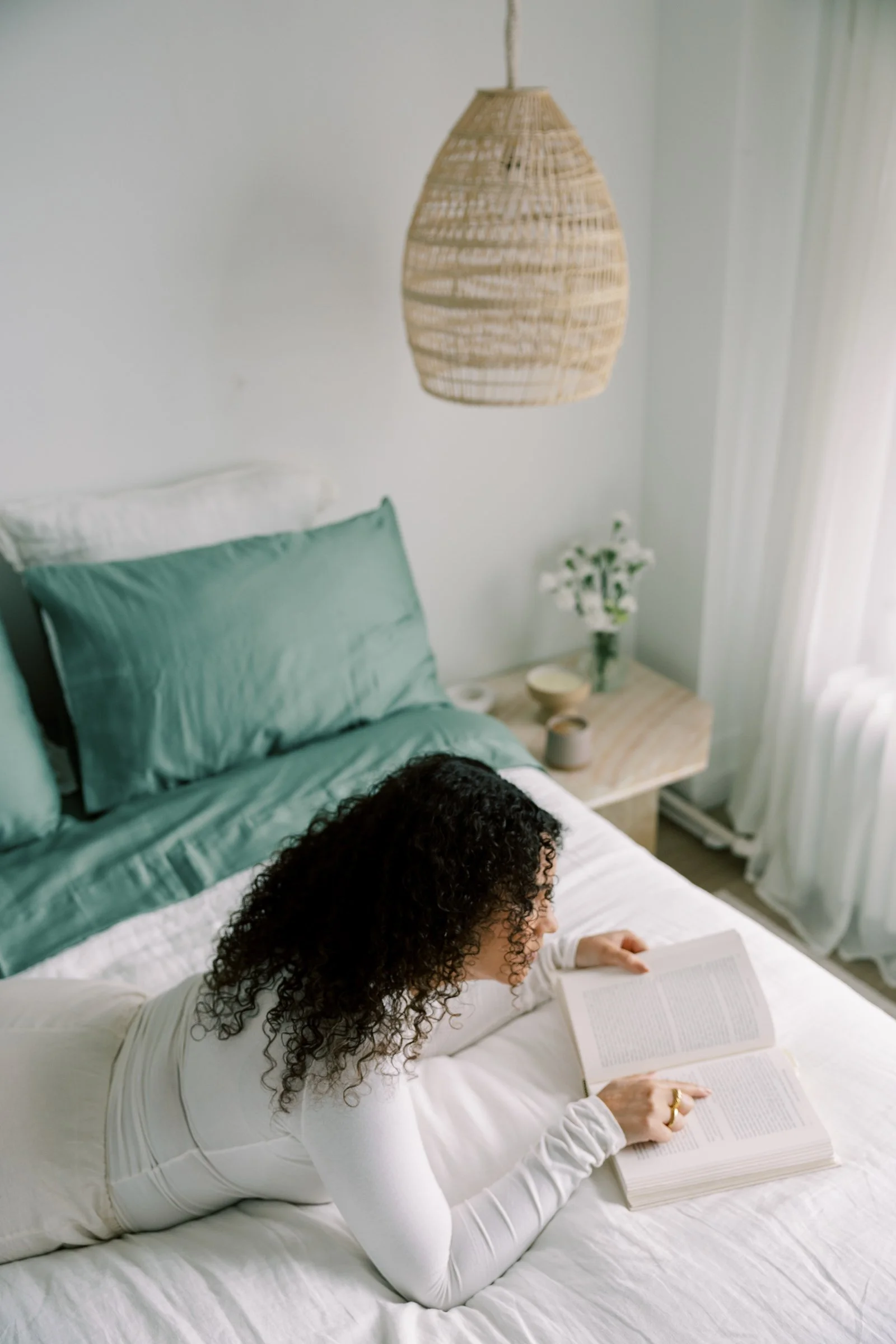 A woman with curly dark hair lying on her stomach on a bed, reading a book in a bright room with white walls, a rattan pendant lamp, green pillows, a small wooden nightstand with a vase of white flowers, and white curtains.
