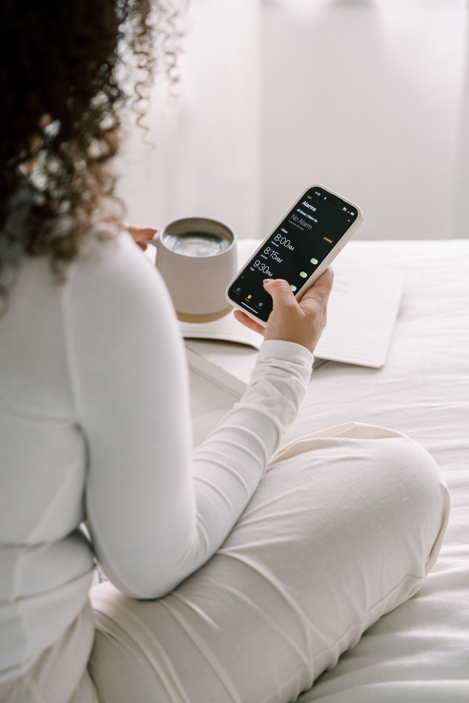 A woman sitting on a bed checking her phone with a wake-up alarms app open, a notebook, a cup of coffee, and a book on the bed.