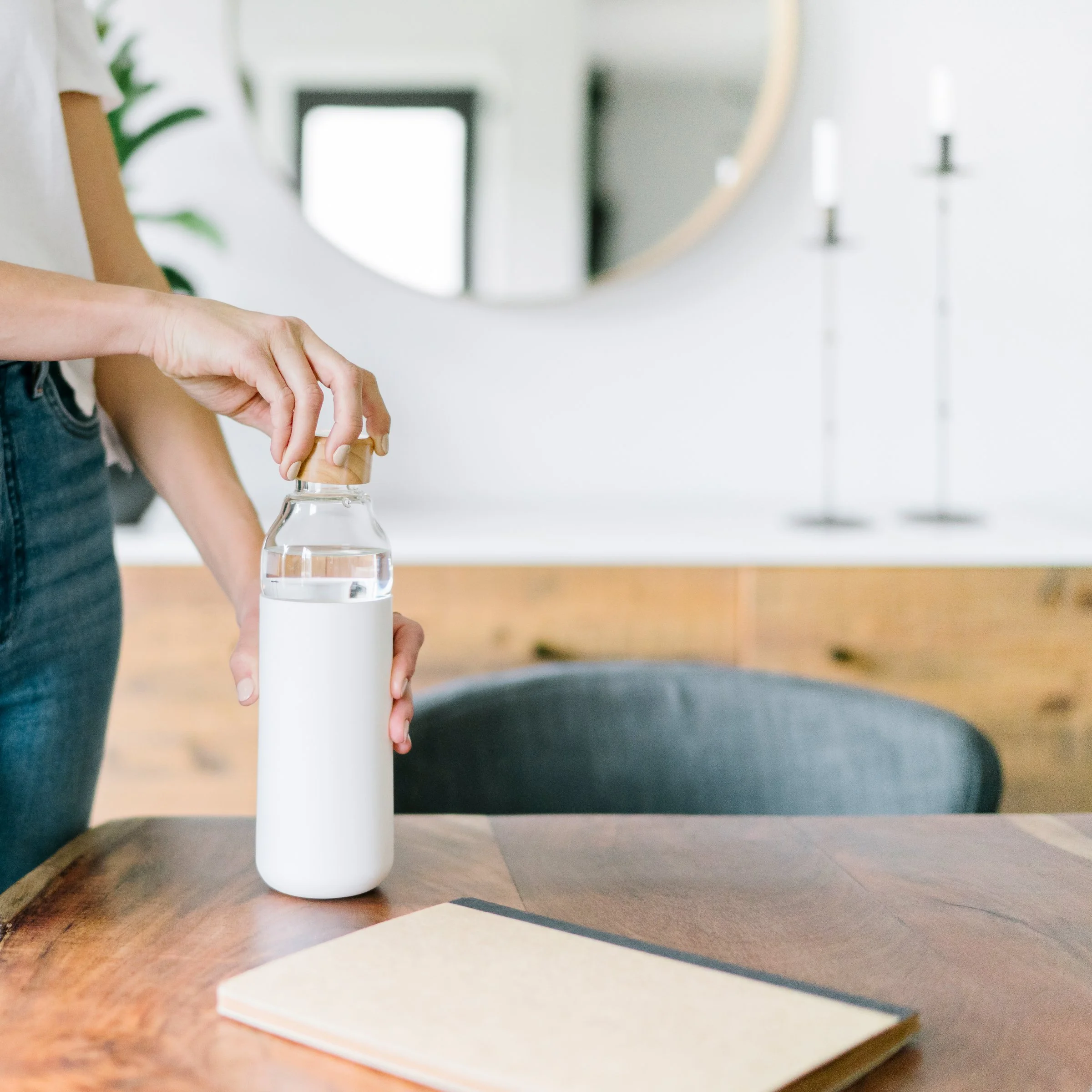 Person opening a glass water bottle with a wooden cap on a wooden table, in a modern home setting.