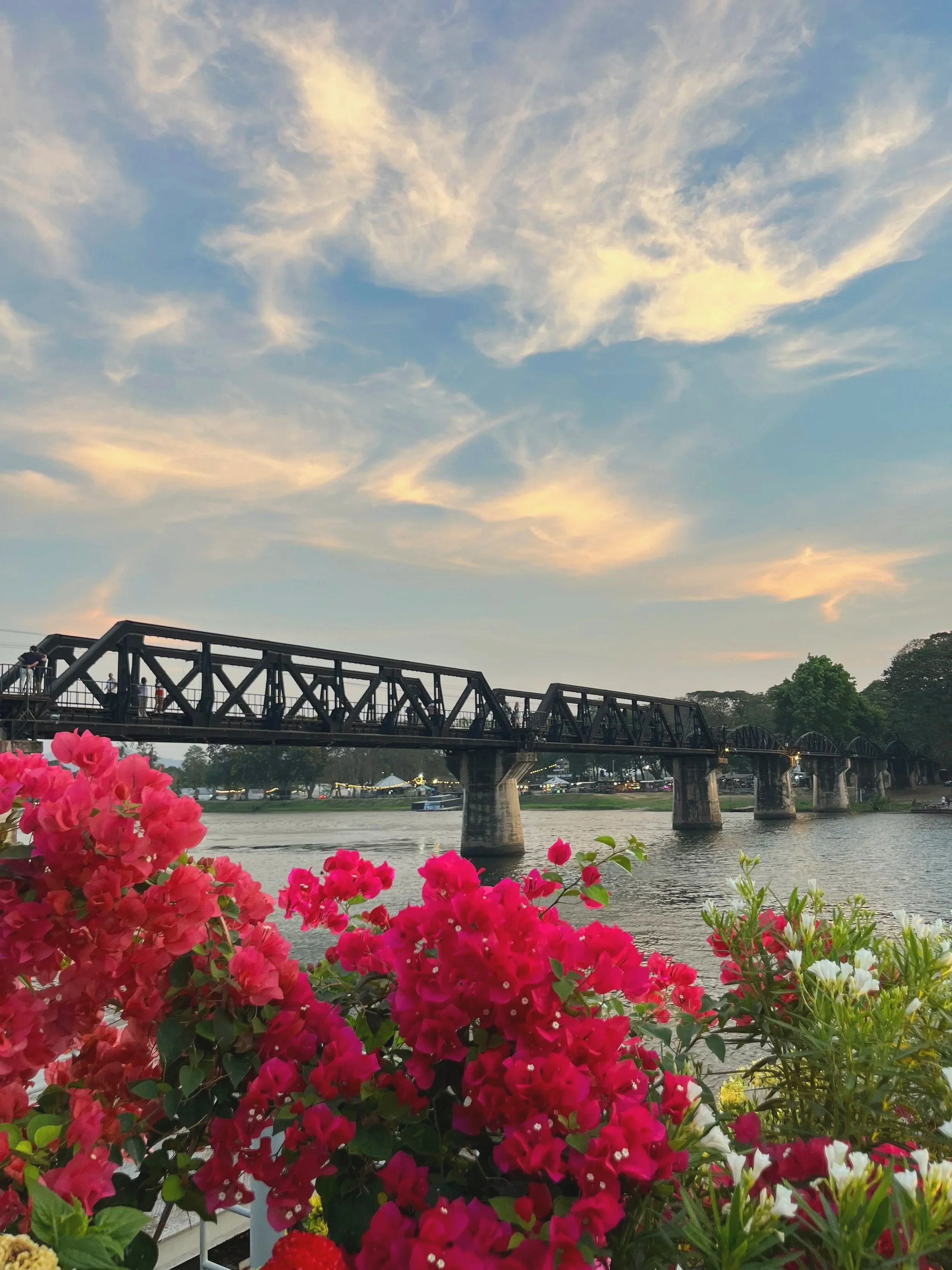 A view of a bridge over a river with colorful flowers in the foreground and a partly cloudy sky.
