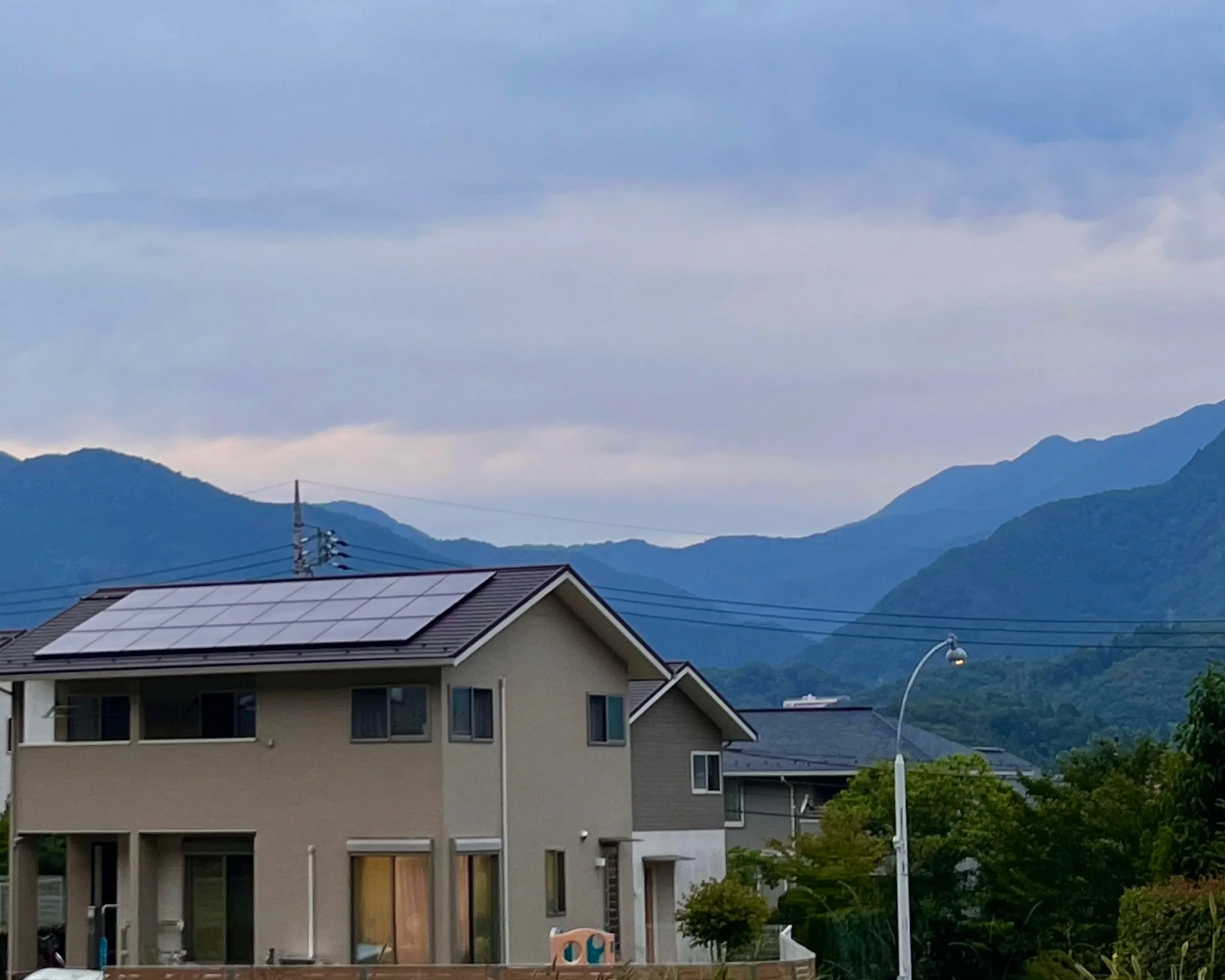 Residential houses with solar panels on the roof, a street lamp, a backdrop of green mountains, and a cloudy sky.