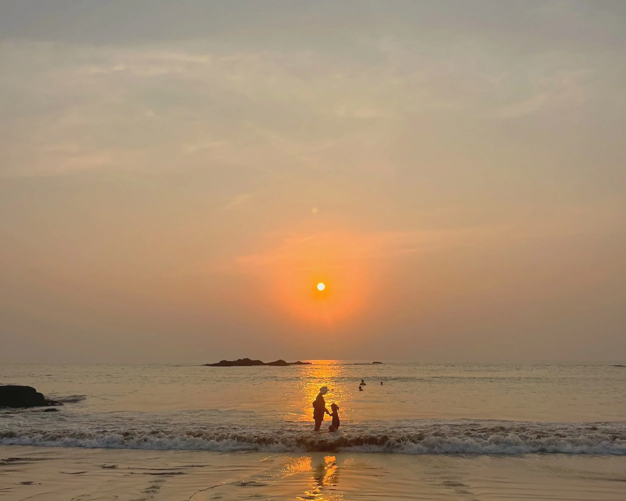 A sunset at the beach with a person and a child standing in the shallow water, surrounded by the warm glow of the setting sun and silhouettes of other swimmers.