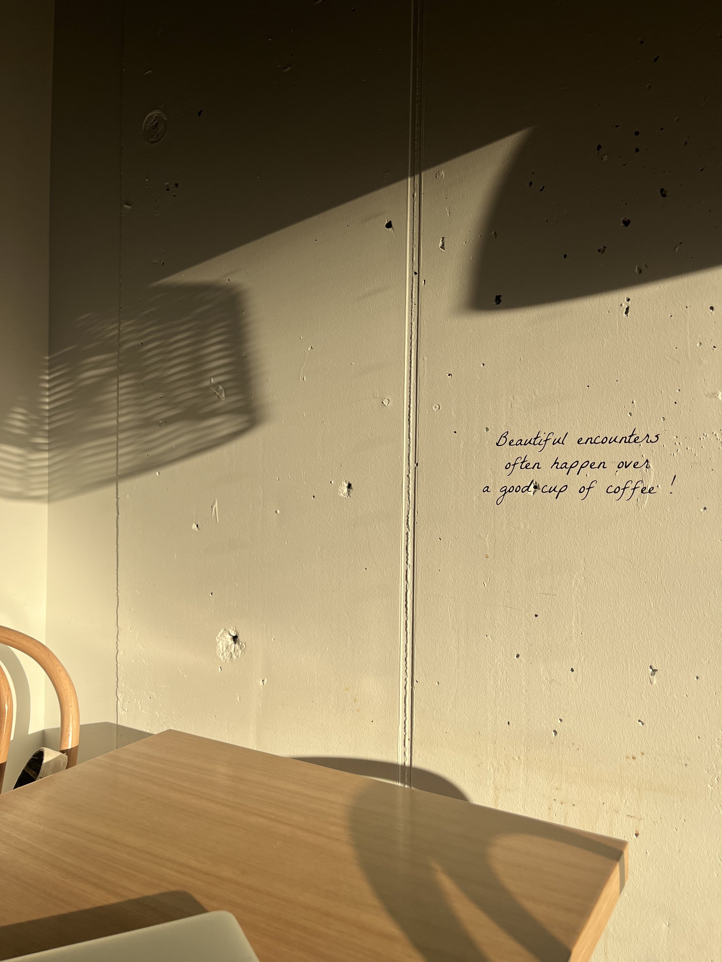 A beige wall with small holes and a handwritten quote that reads, "Beautiful encounters often happen over a good cup of coffee!" Shadows cast by a window or light fixture fall across the wall and table.