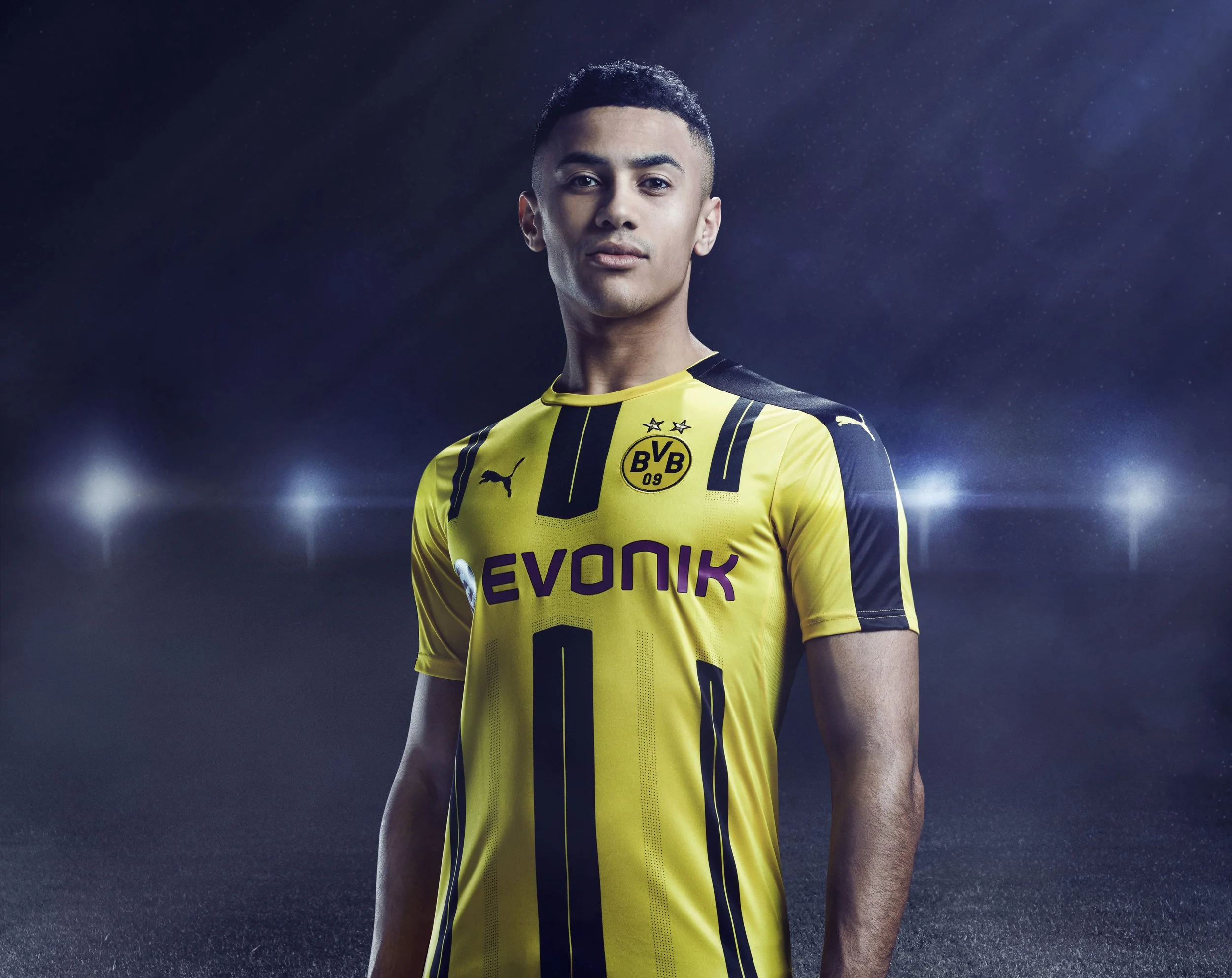 A young man in a yellow and black Borussia Dortmund soccer jersey posing against a dark, starry background with stadium lights.