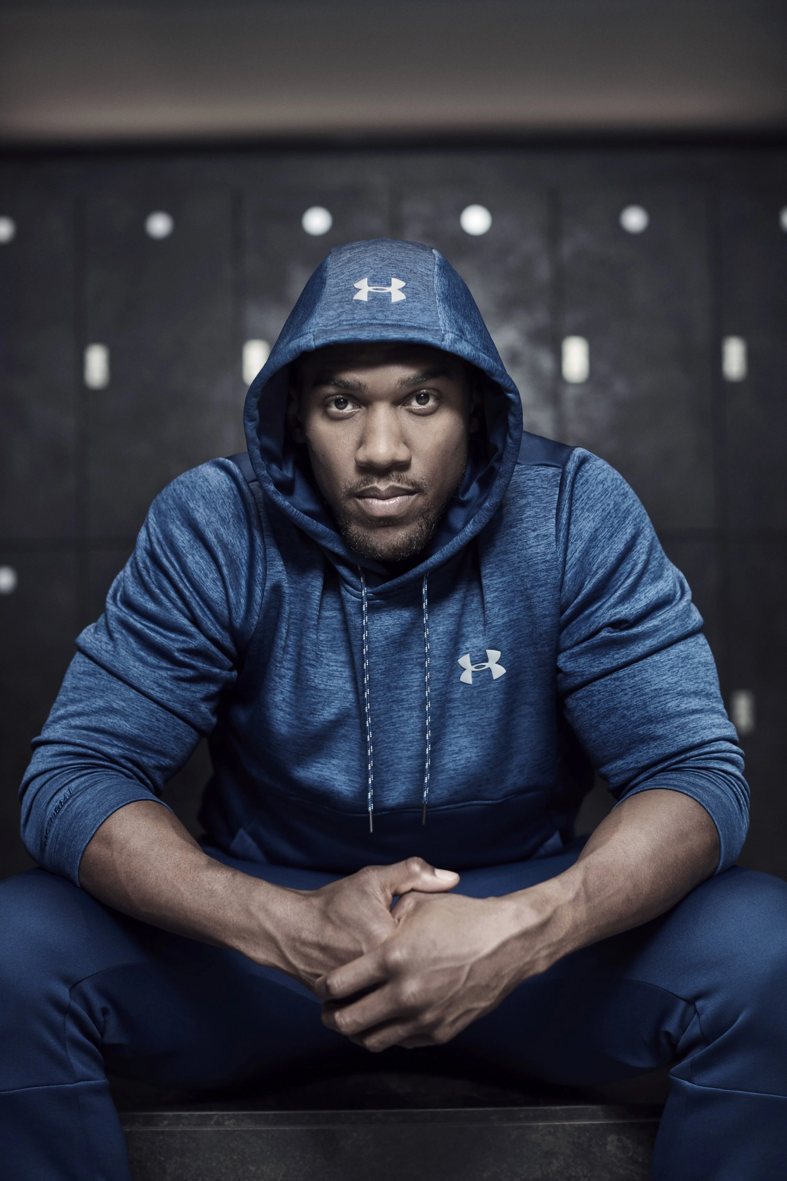 Anthony Oluwafemi Olaseni "AJ" Joshua is a British professional boxer, photography for JD set in a changing rooms, wearing a blue Under Armour hoodie sitting with his arms crossed, looking directly at the camera in a locker room.