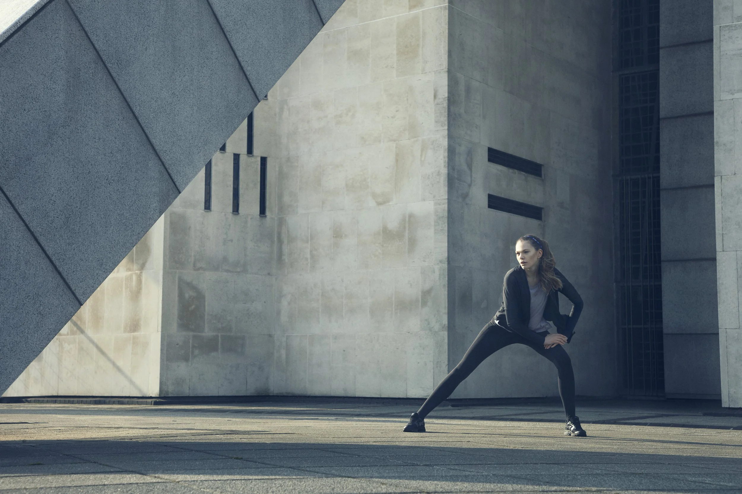 A woman with long hair in athletic clothing performing a stretching exercise in front of modern concrete building with geometric shapes and windows.