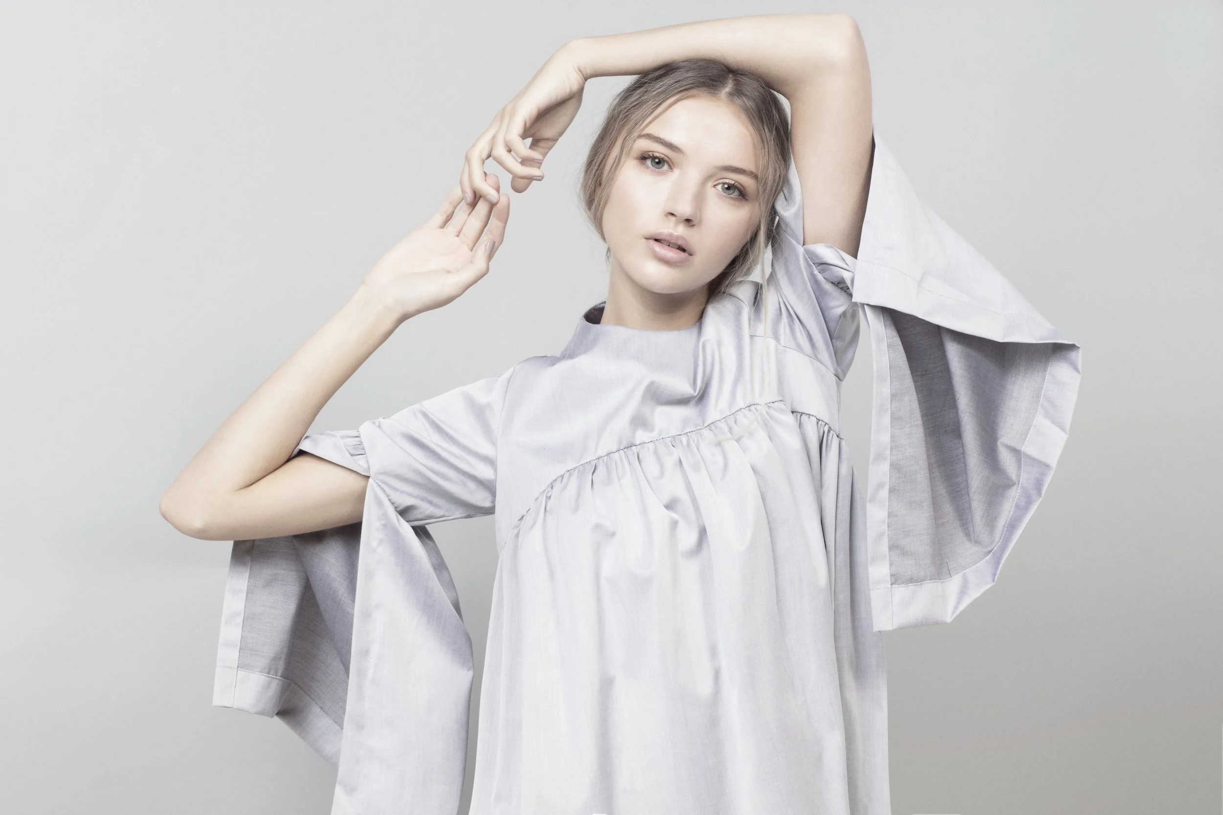 A young woman with styled short hair and fair skin posing against a plain gray background. She wears a loose, light-colored dress with wide, short sleeves. She has one arm raised above her head and the other hand gently touching her face, with a neut