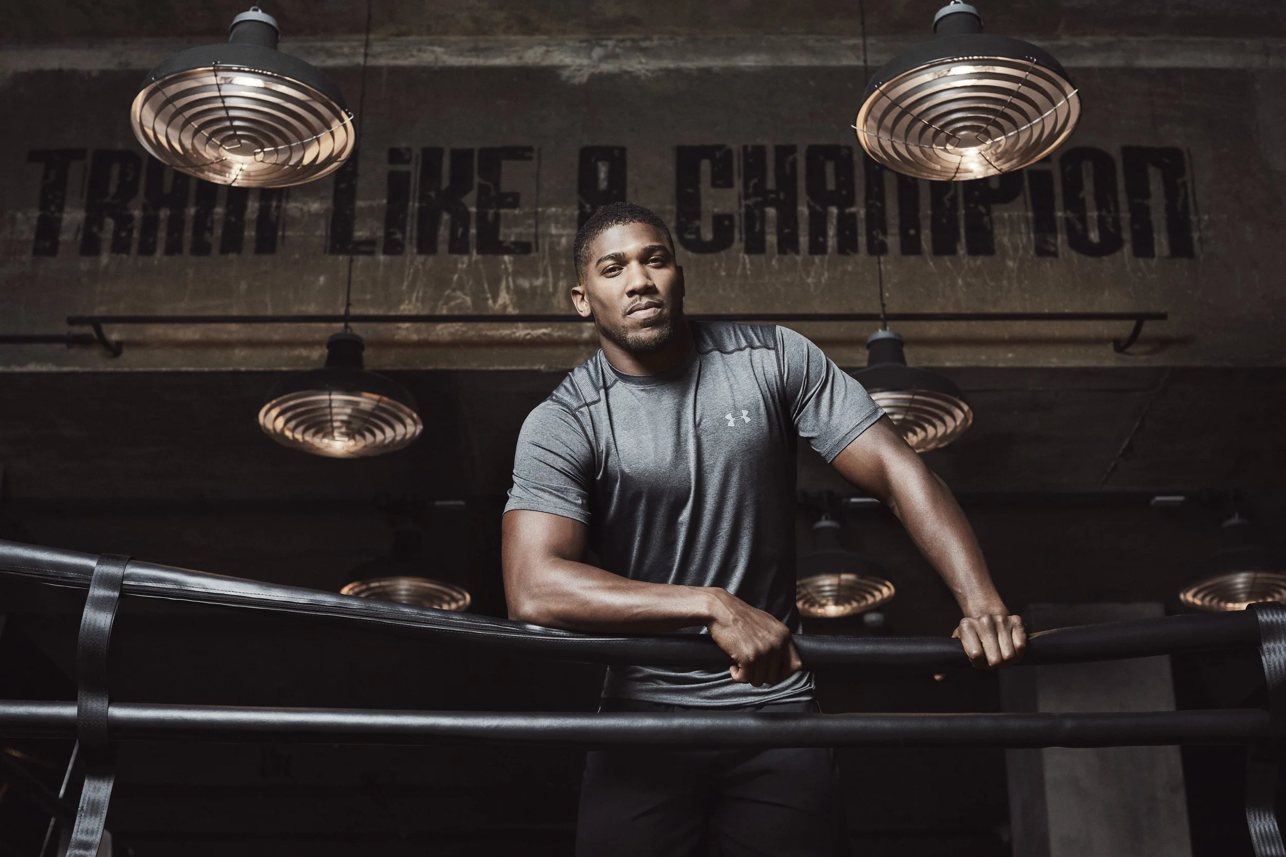 Anthony Oluwafemi Olaseni "AJ" Joshua is a British professional boxer,  photography for JD set in a boxing ring, in a gray athletic shirt workout at a gym with industrial lighting and a wall that reads 'THE FURTHER YOU CHALLENGE'