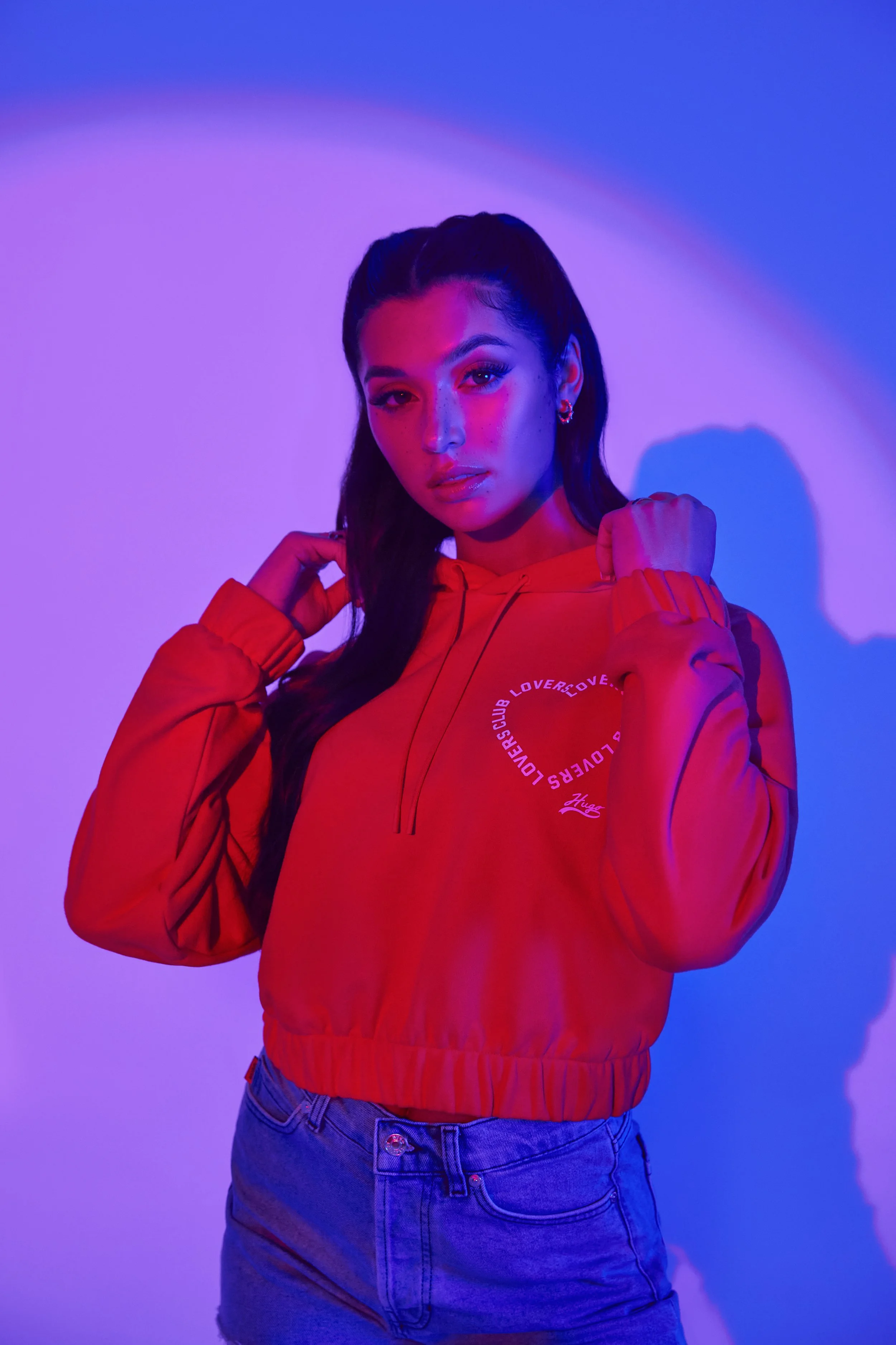 A young woman with long dark hair styled in a high ponytail, wearing a red hooded jacket with white circular text design on the chest, standing against a background with purple and blue lighting.