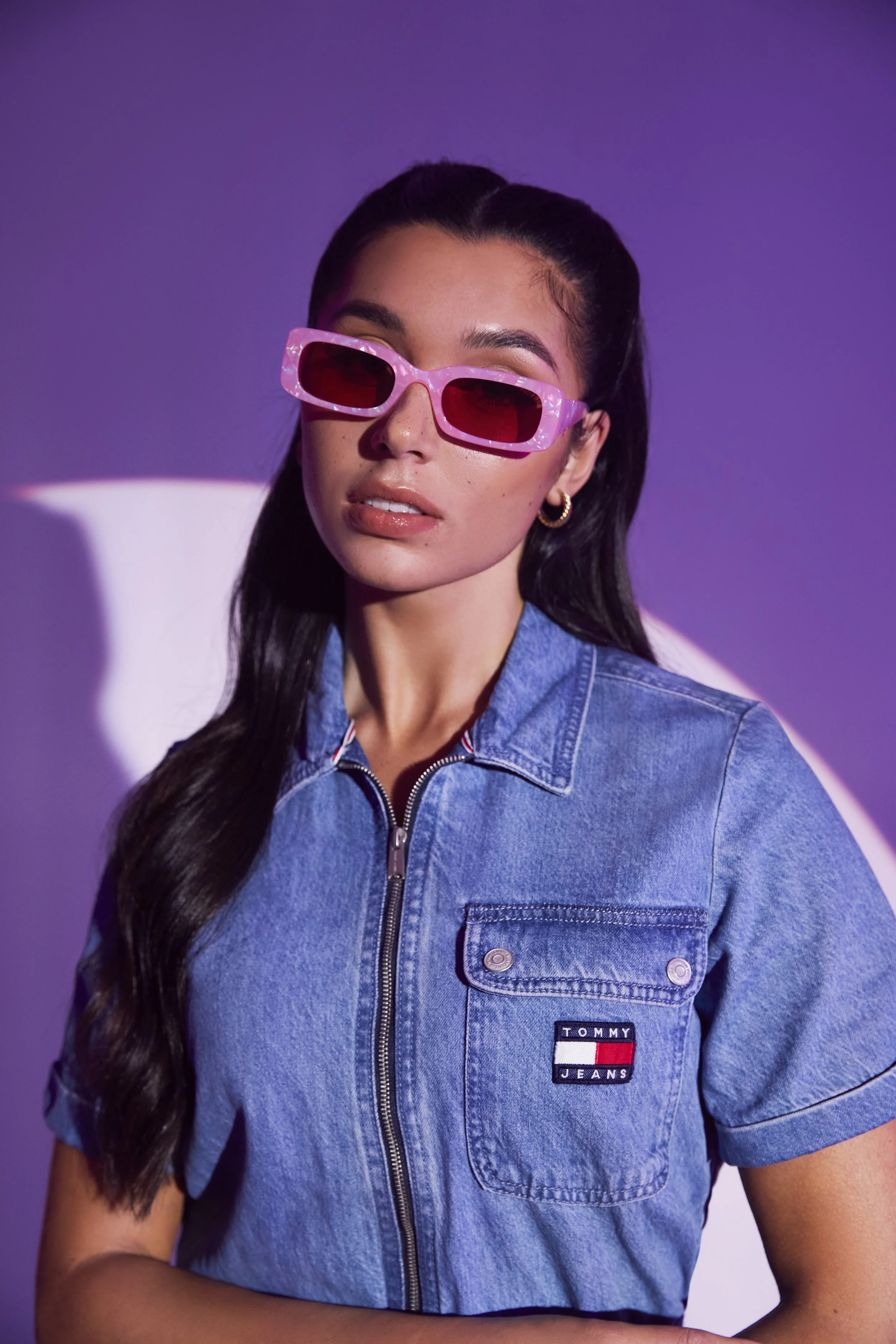 A woman wearing pink sunglasses and a denim jacket with a Tommy Jeans logo stands against a purple background.
