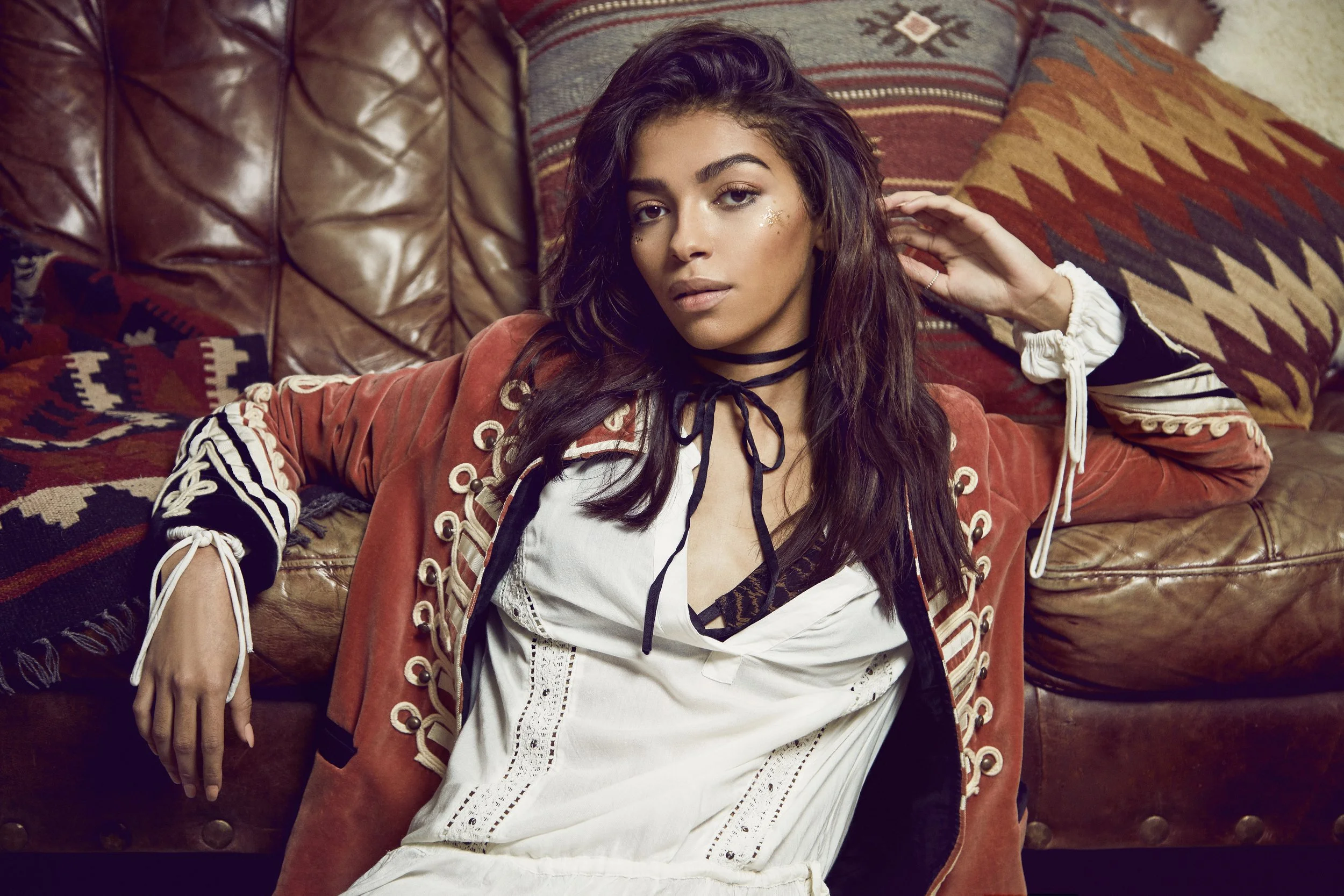 A young woman with long dark hair and makeup, including glitter, resting against a leather couch with patterned pillows, wearing a white dress and a rust-colored jacket with decorative buttons and lacing.