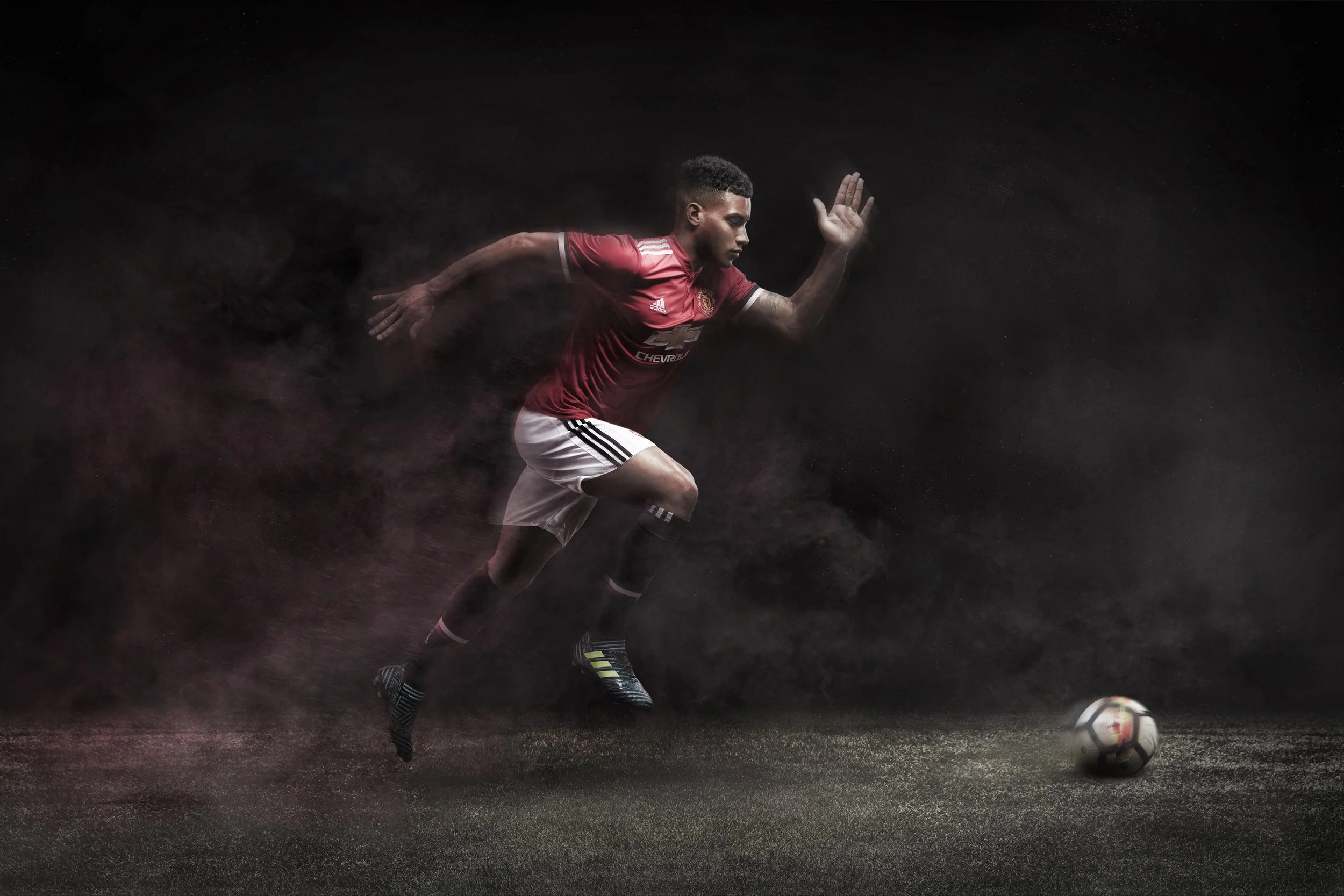 Manchester United photoshoot, A male athlete in red and white sportswear is mid-action running after a soccer ball on a dark background, with motion effects and dust or particles around him.