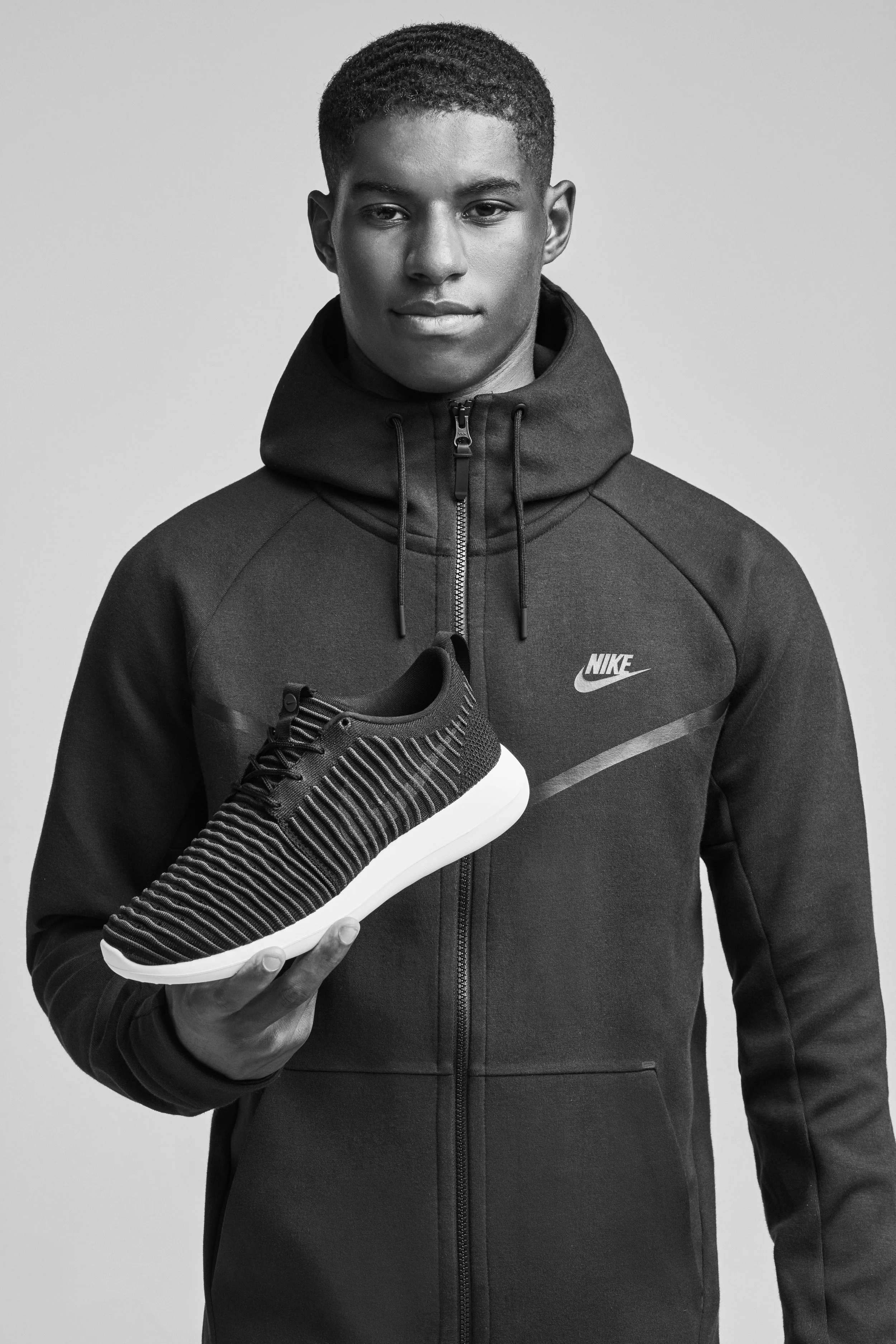 Marcus Rashford is an English professional footballer, holding a black and white athletic shoe, wearing a Nike sports jacket, in a black-and-white portrait against a plain background.