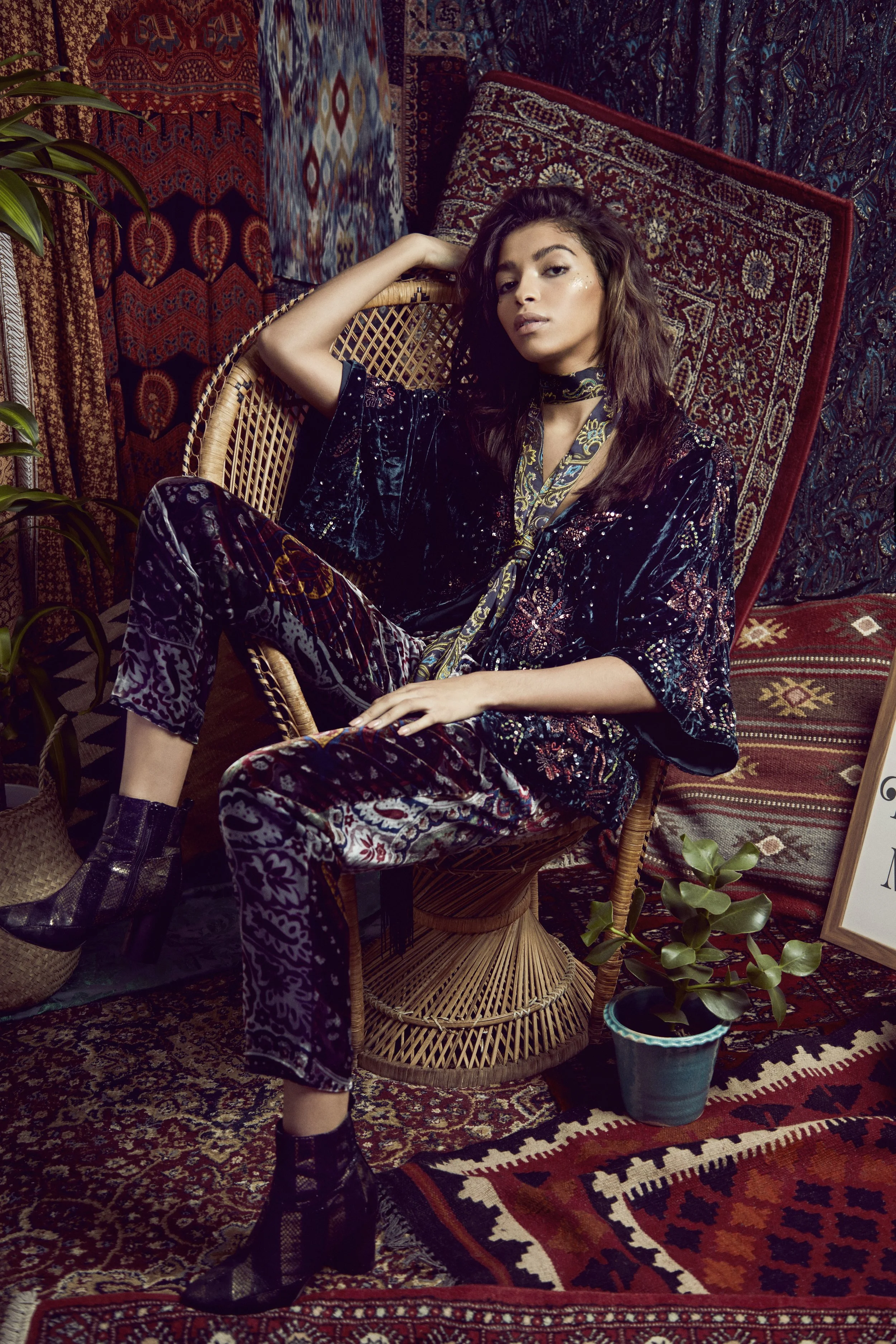 A woman with long dark hair wearing a dark velvet floral embroidered jacket and patterned pants, sitting in a wicker chair in a room decorated with various colorful Oriental rugs and a green potted plant.