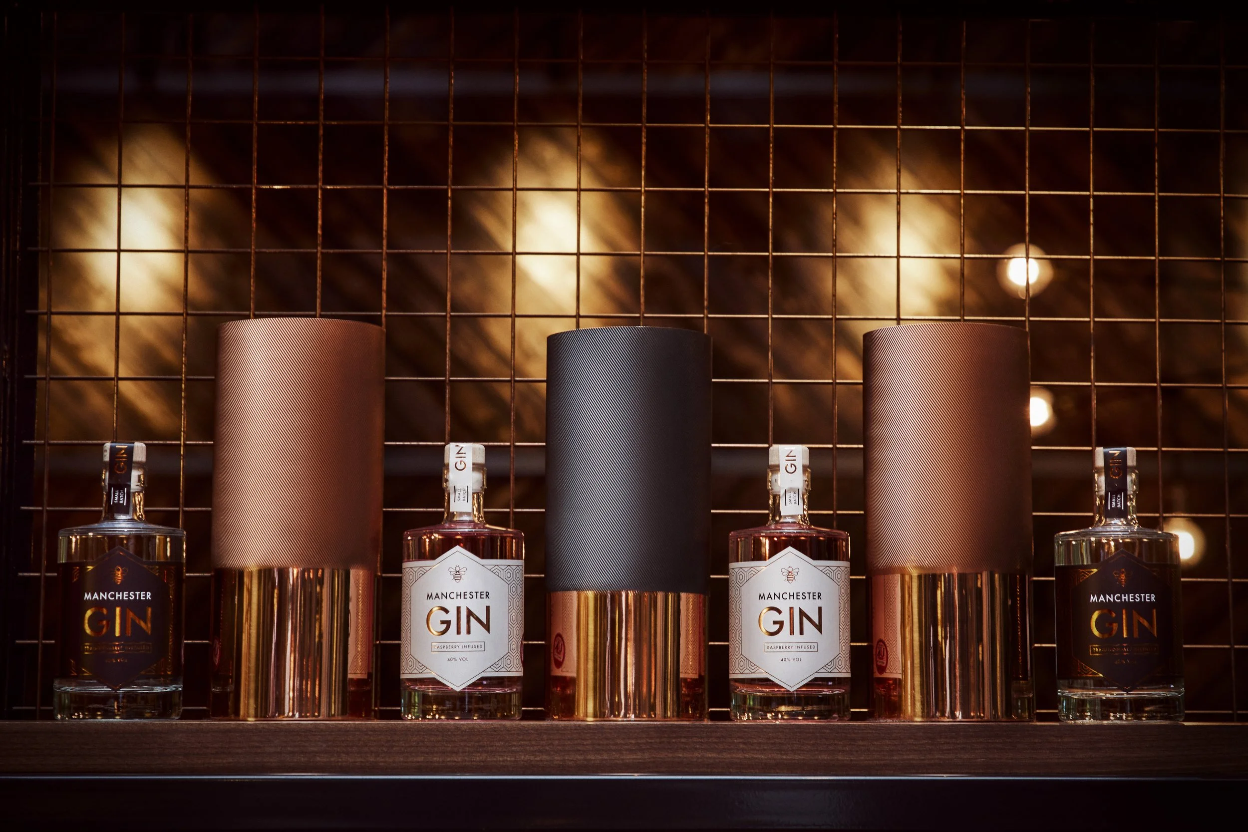 Several bottles of Manchester gin displayed on a shelf, with three cylindrical containers behind them, set against a metal grid background with warm lighting.