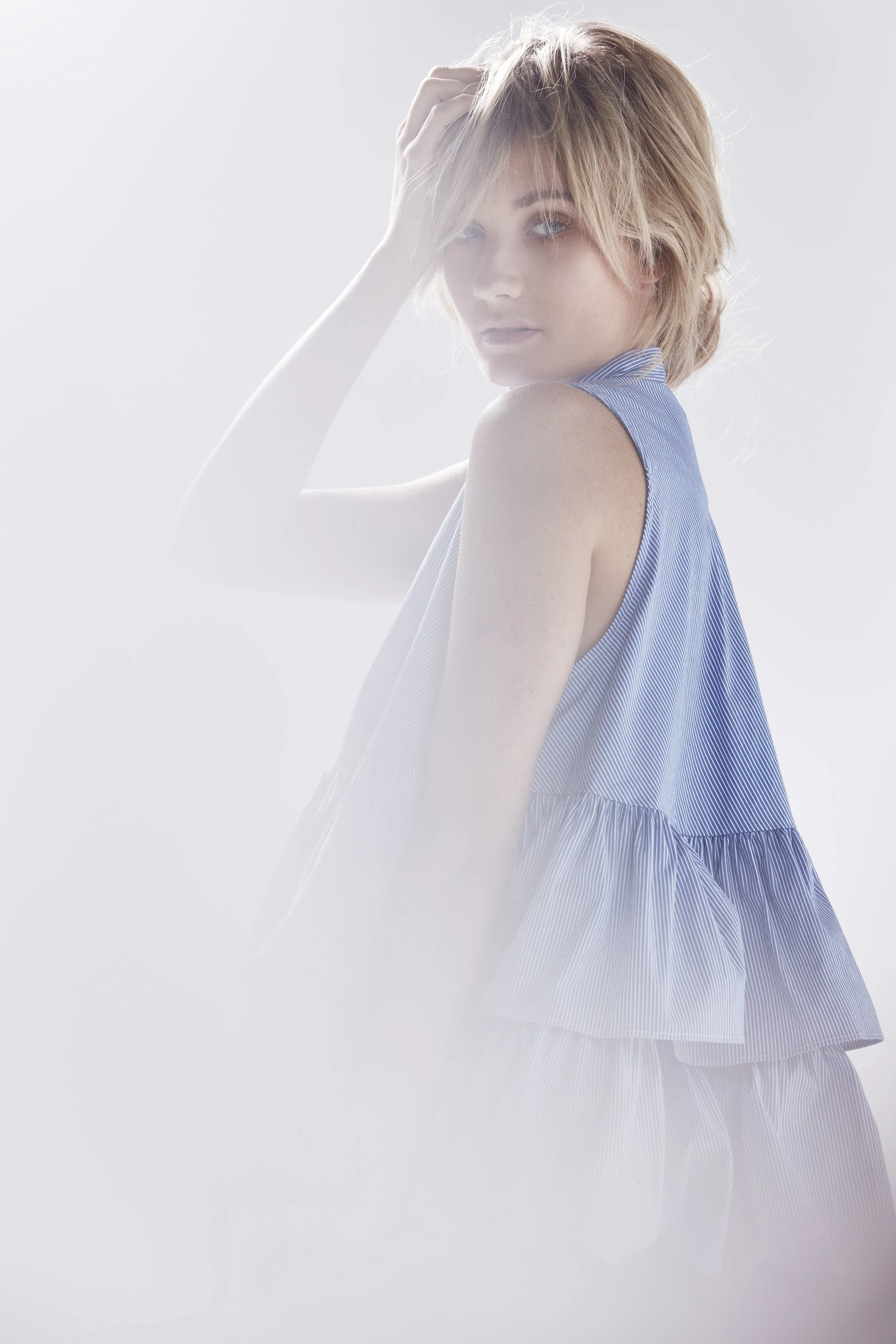 A young woman with short, wavy blonde hair wearing a sleeveless, blue, striped dress, looking over her shoulder and touching her hair against a plain white background.