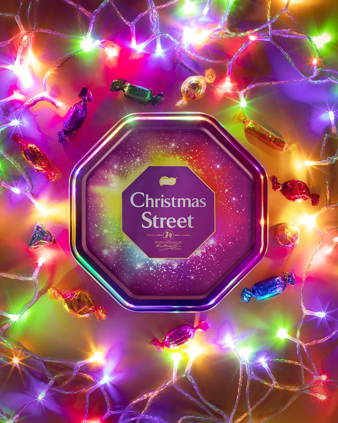 A purple tin of Nestlé Christmas Street chocolates surrounded by colorful string lights and wrapped candies.