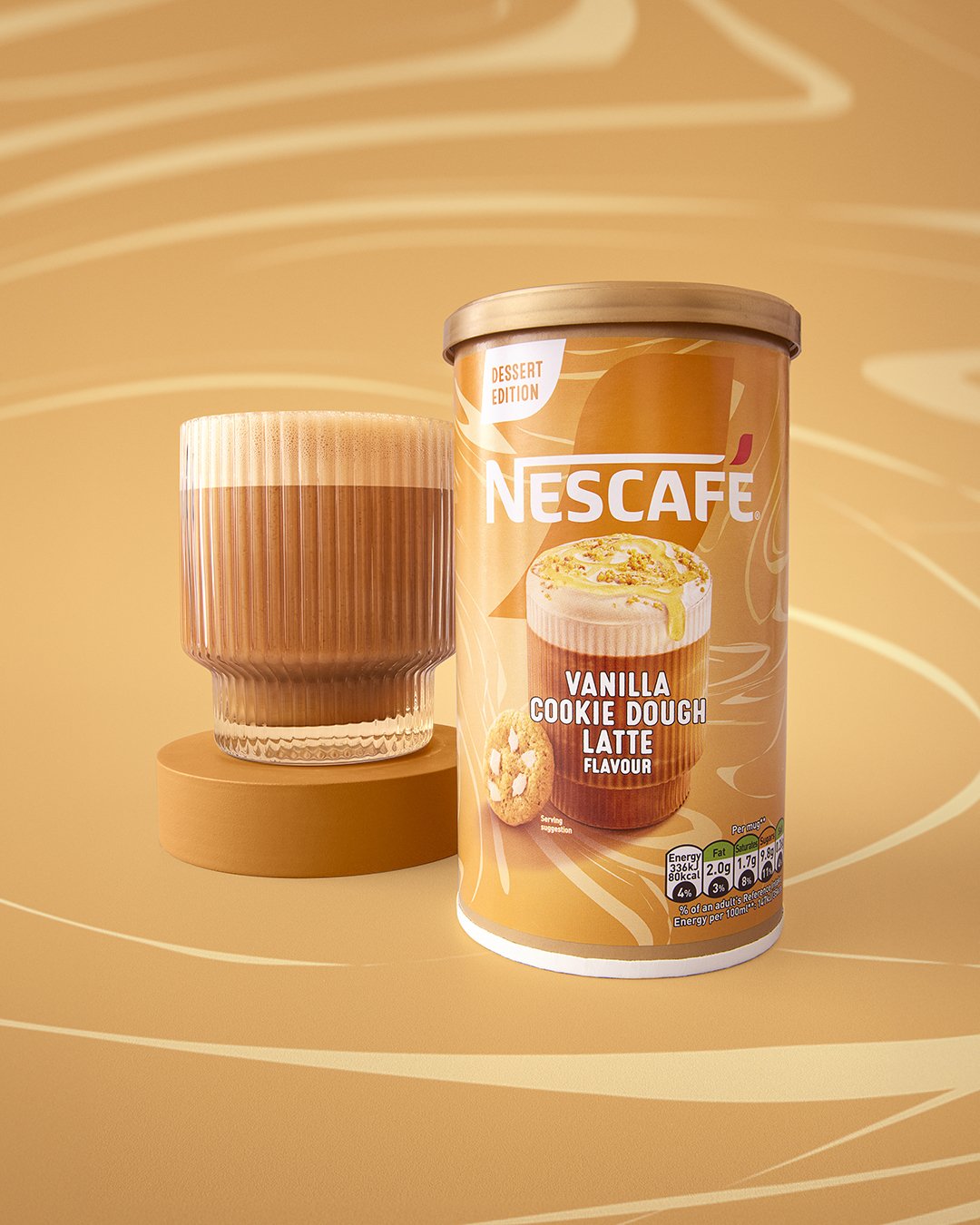A canister of NESCAFÉ Vanilla Cookie Dough Latte flavor and a glass of the latte drink on a round beige platform, set against a matching beige background with abstract wavy lines.