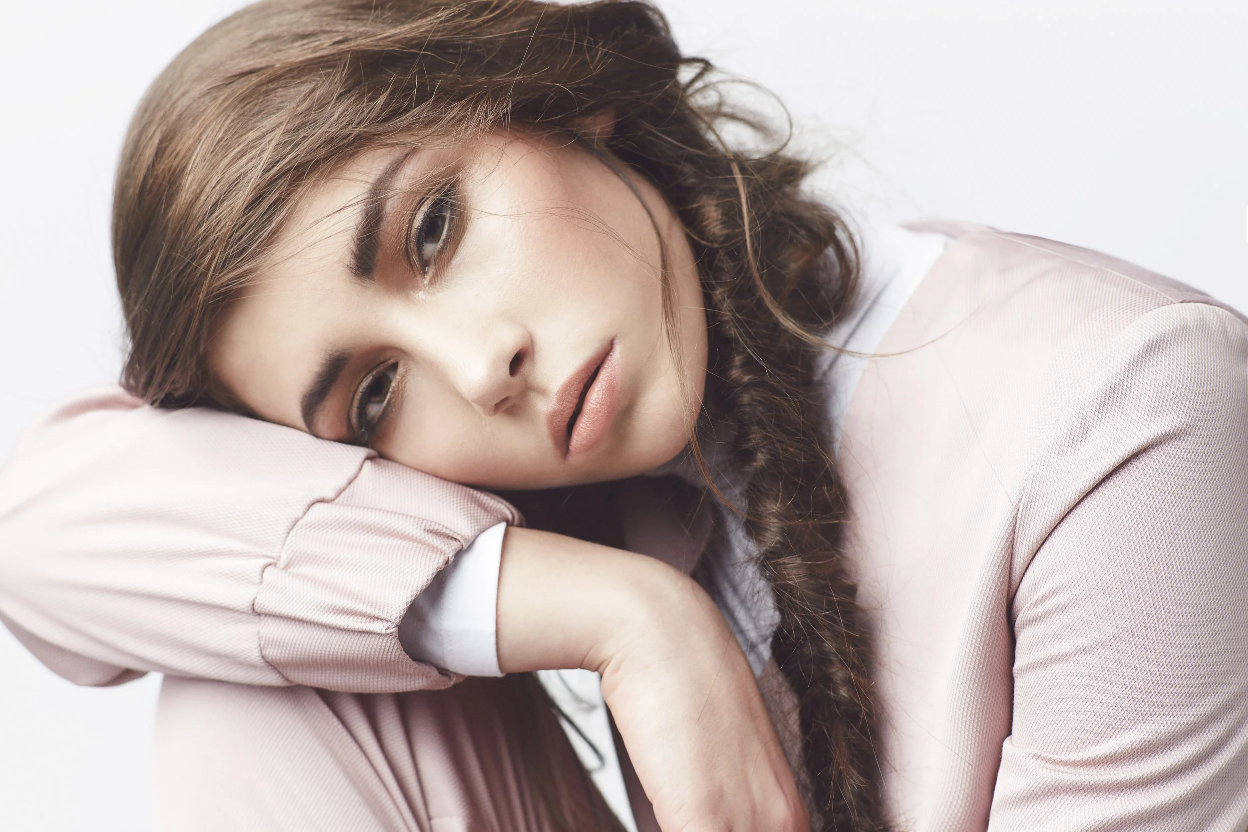 A woman with curly brown hair, resting her head on her arm, wearing a light pink long-sleeve shirt, with a neutral expression and a white background.