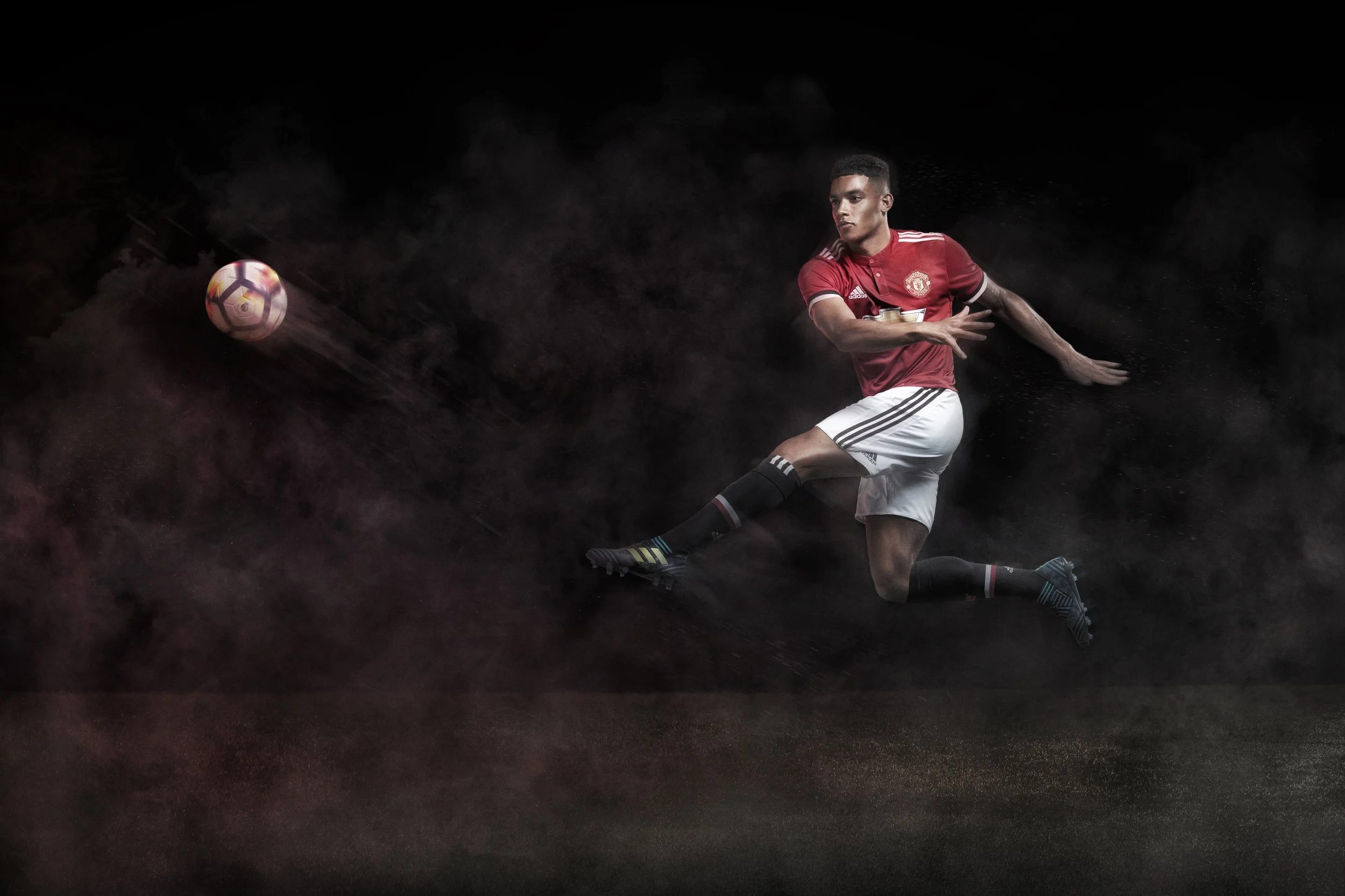 Manchester United photoshoot, A male soccer player in a red jersey and white shorts kicking a soccer ball against a dark background.