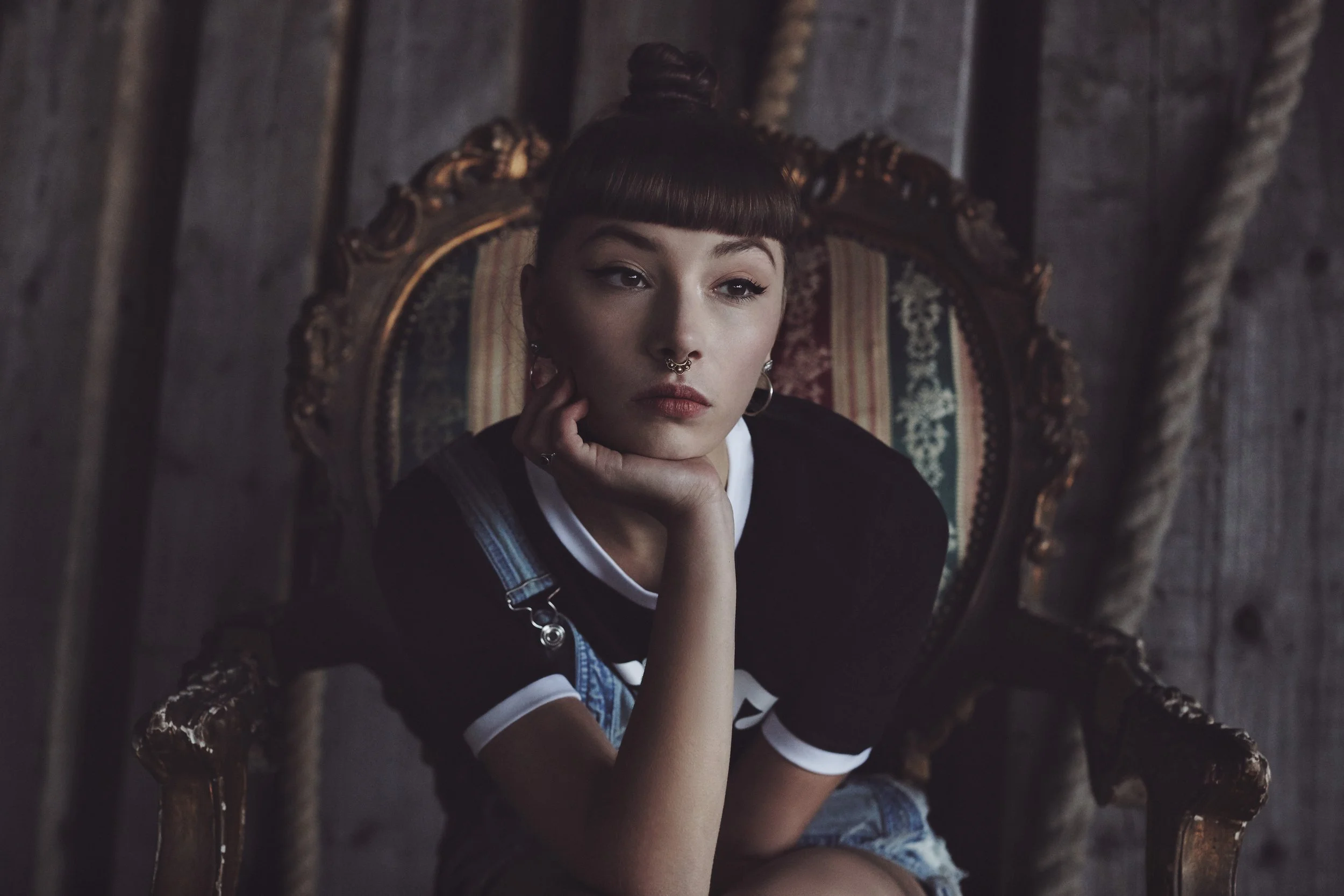 A young woman with a septum piercing and dark hair in a bun, sitting on an ornate wooden chair with a patterned backrest, in a dimly lit room with wooden walls.