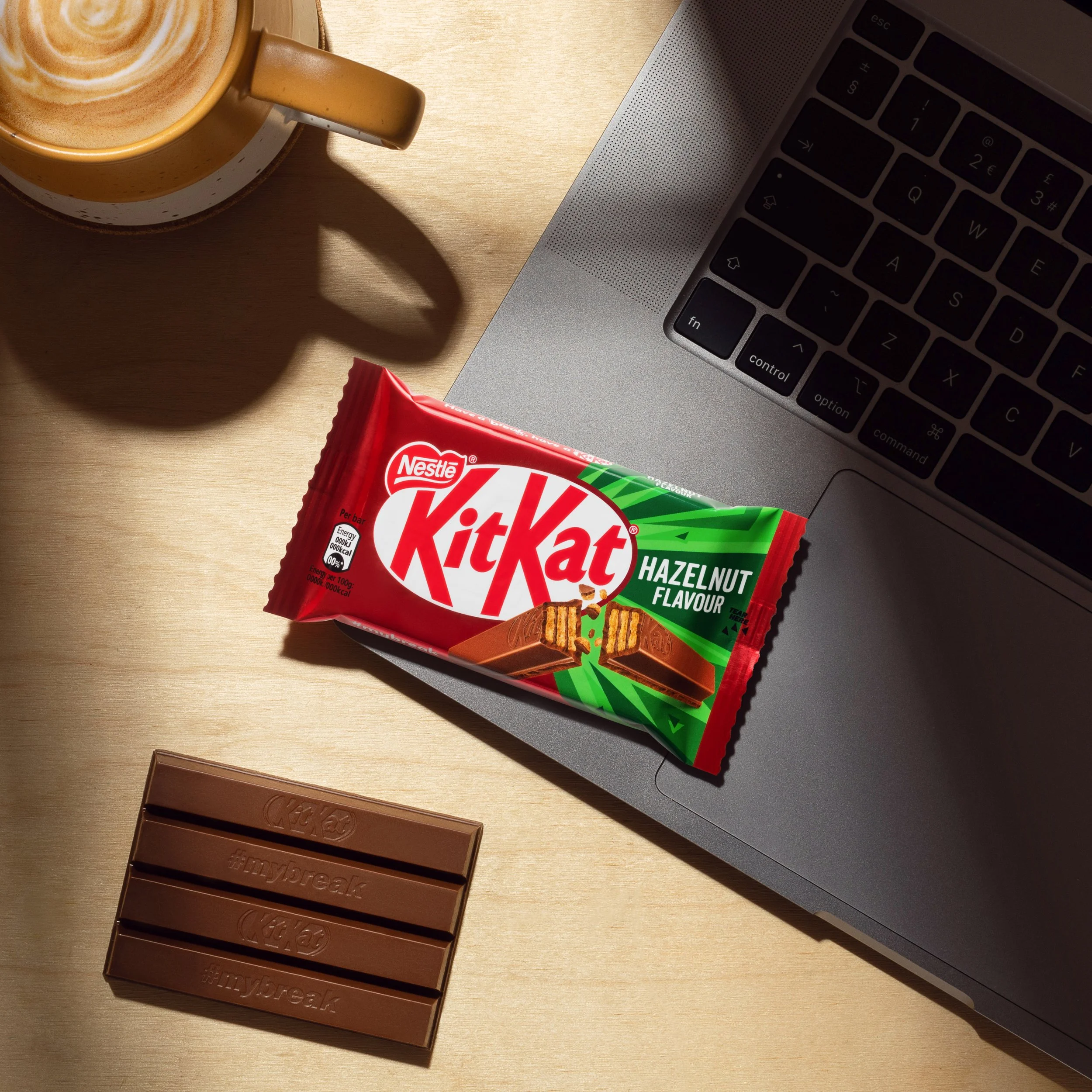 A laptop keyboard, a bar of milk chocolate, a bar of milk chocolate with hashtag #mybreak, a cup of coffee with latte art on a saucer, and a package of Kit Kat Hazelnut Flavor on a wooden surface.