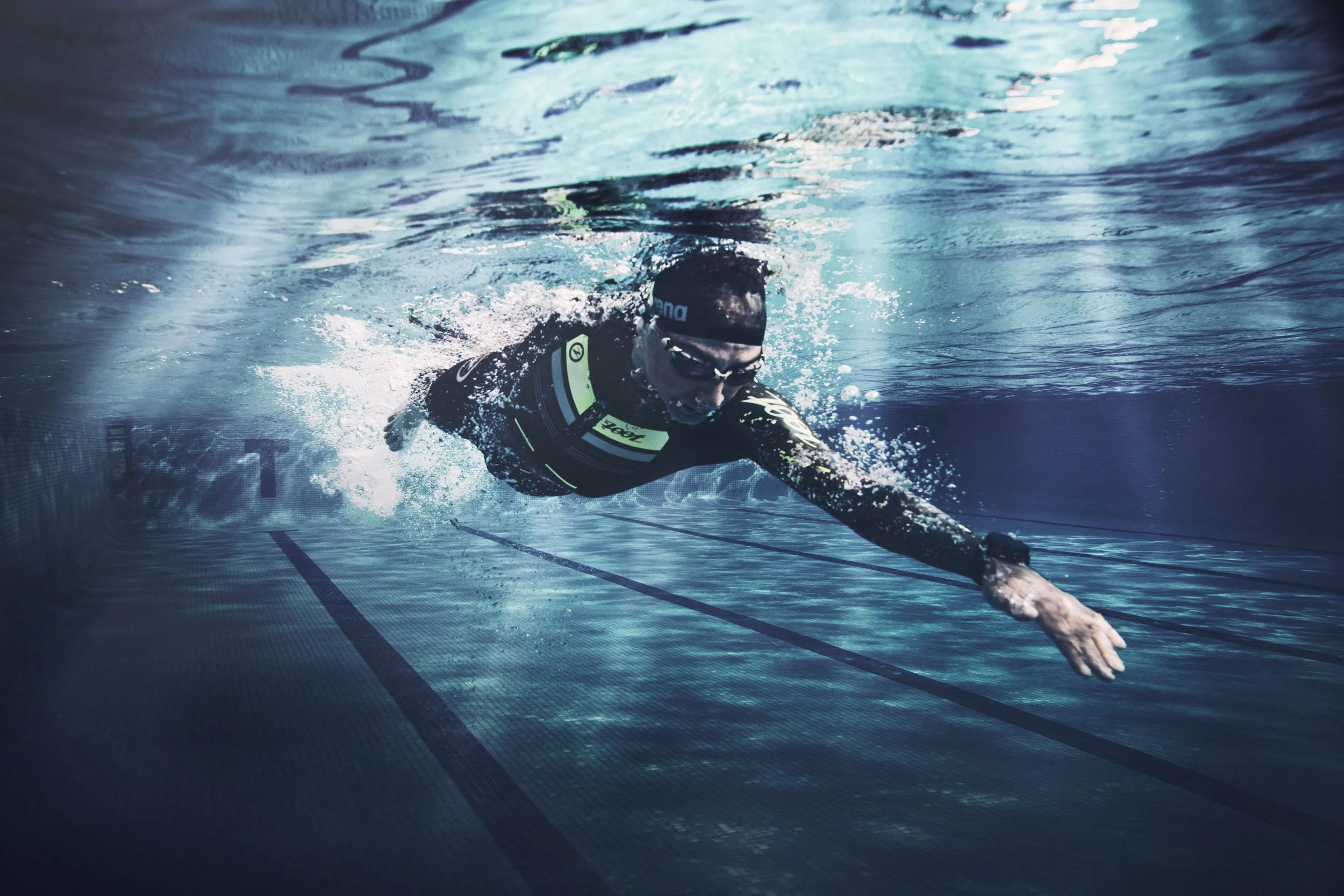 Swimmer in an underwater pool, wearing a swim cap and goggles, extending their arm forward during a swim stroke.