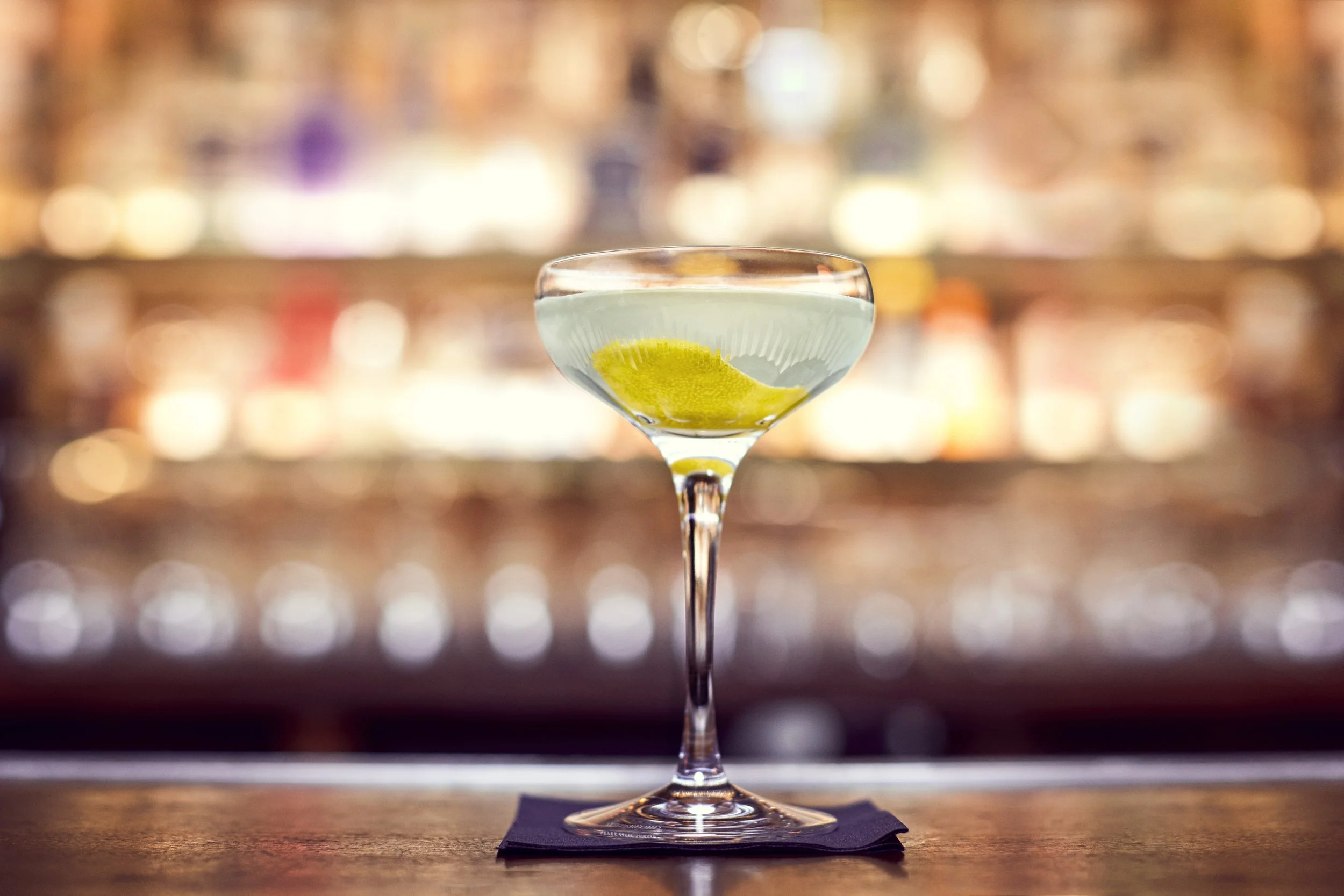 A cocktail glass on a black napkin placed on a wooden bar counter, filled with a clear drink and a lime wedge garnish, with blurred multicolored lights in the background.