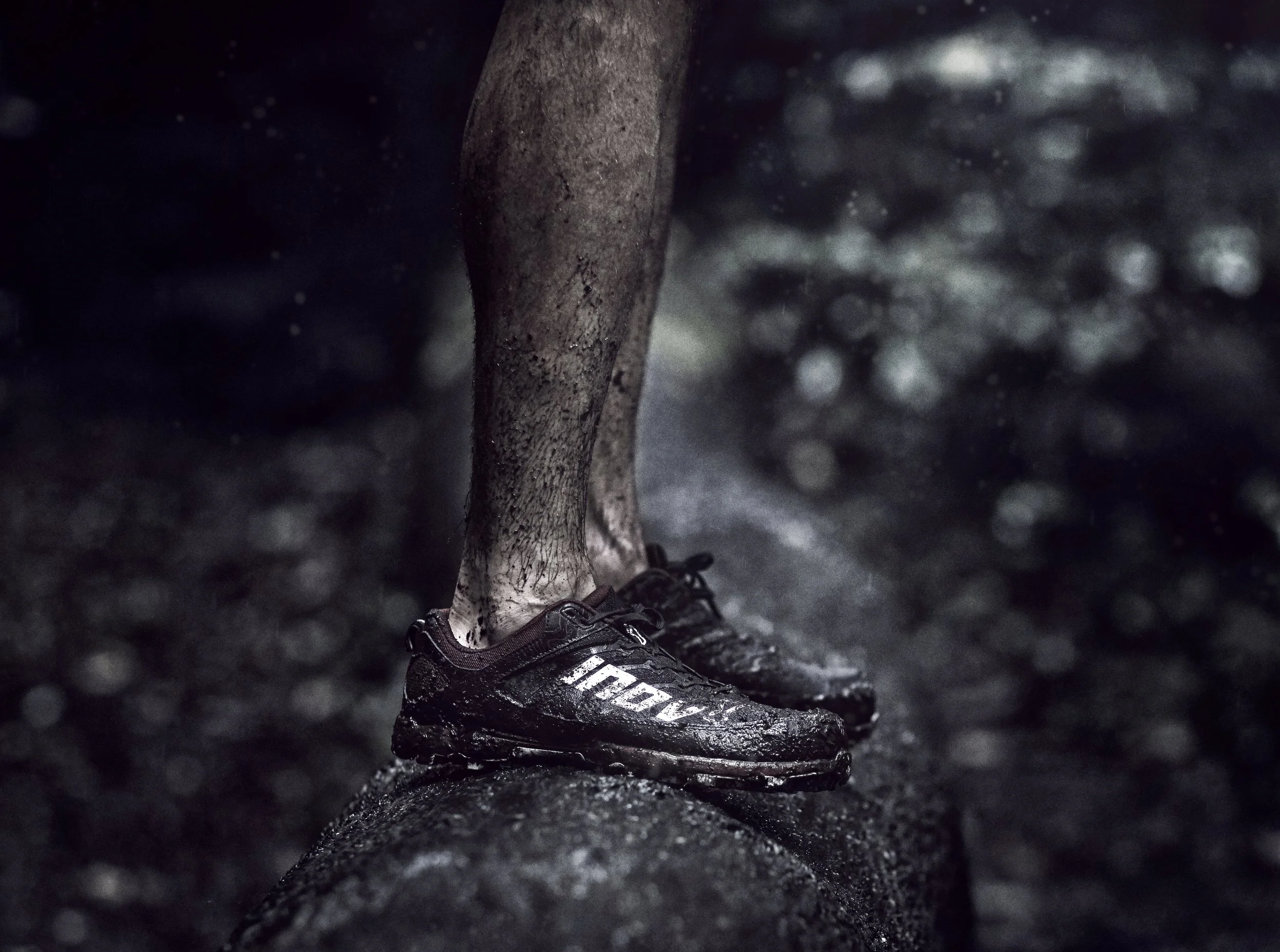 Close-up of a cyclist's foot in a black and white athletic shoe, clipped into a pedal, on a dark, wet trail or road surface.
