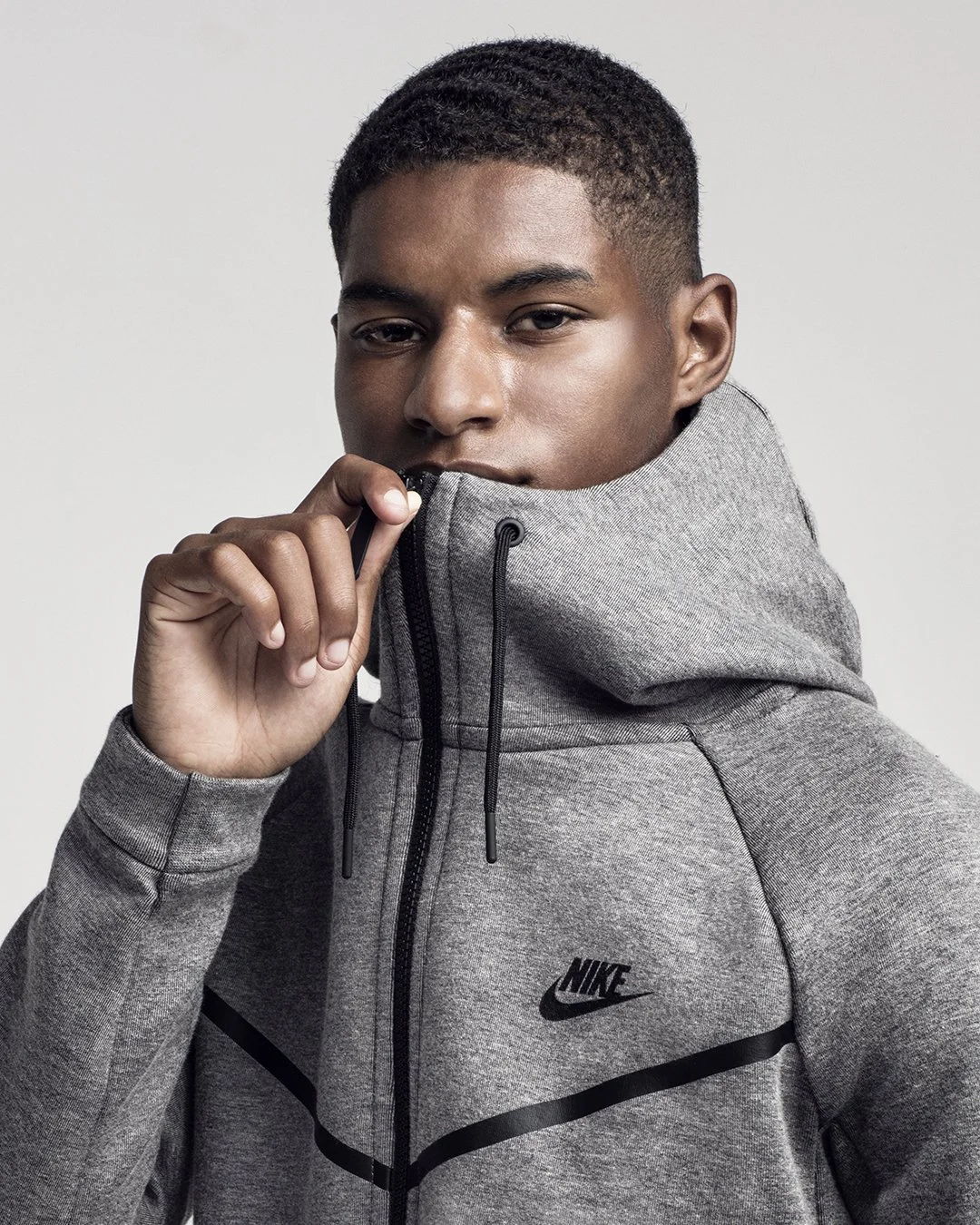 Marcus Rashford is an English professional footballer, wearing a gray Nike zip-up hoodie with the hood up, holding the zipper near his mouth, looking at the camera.