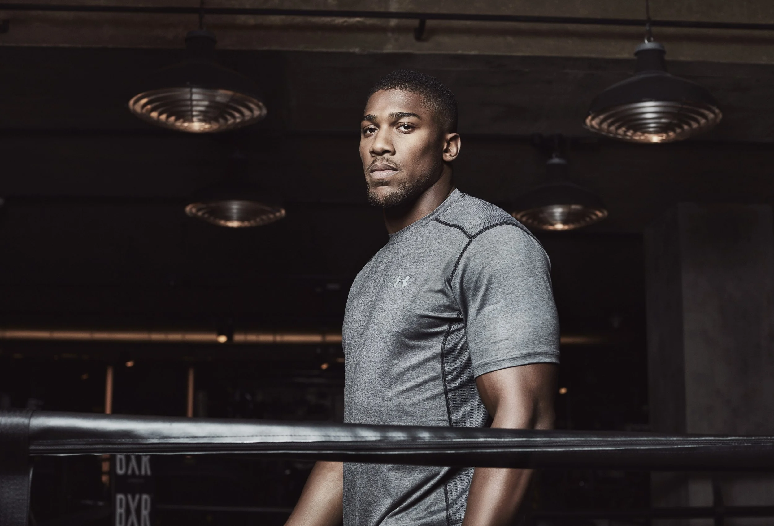 Anthony Oluwafemi Olaseni "AJ" Joshua is a British professional boxer, photography for JD, wearing a gray Under Armour athletic shirt standing in a dark indoor gym with industrial-style ceiling fans.