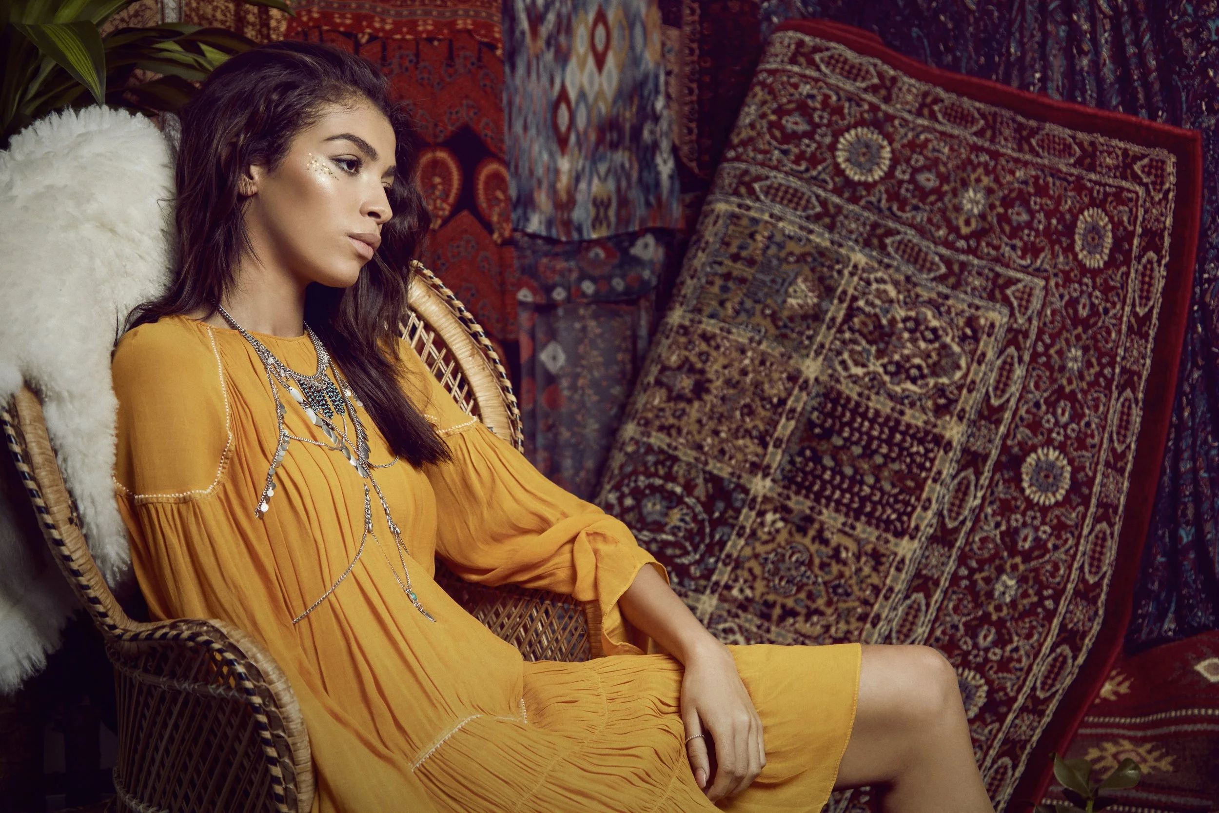 A woman with dark hair and glowing skin resting in a wicker chair, wearing a yellow dress and layered silver necklaces, surrounded by patterned rugs.