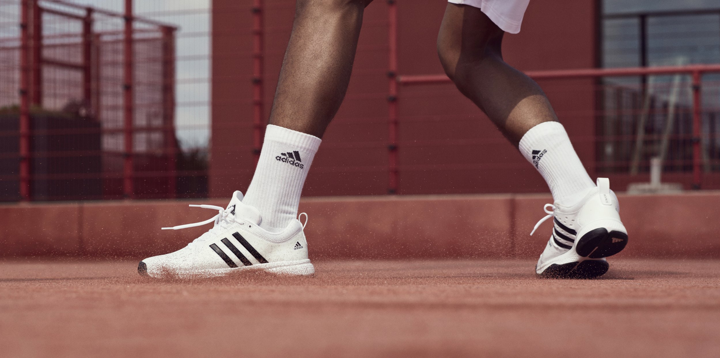 Close-up of a person wearing white Adidas sneakers and white socks with black Adidas logo, standing on a red outdoor court, with a red fence in the background.