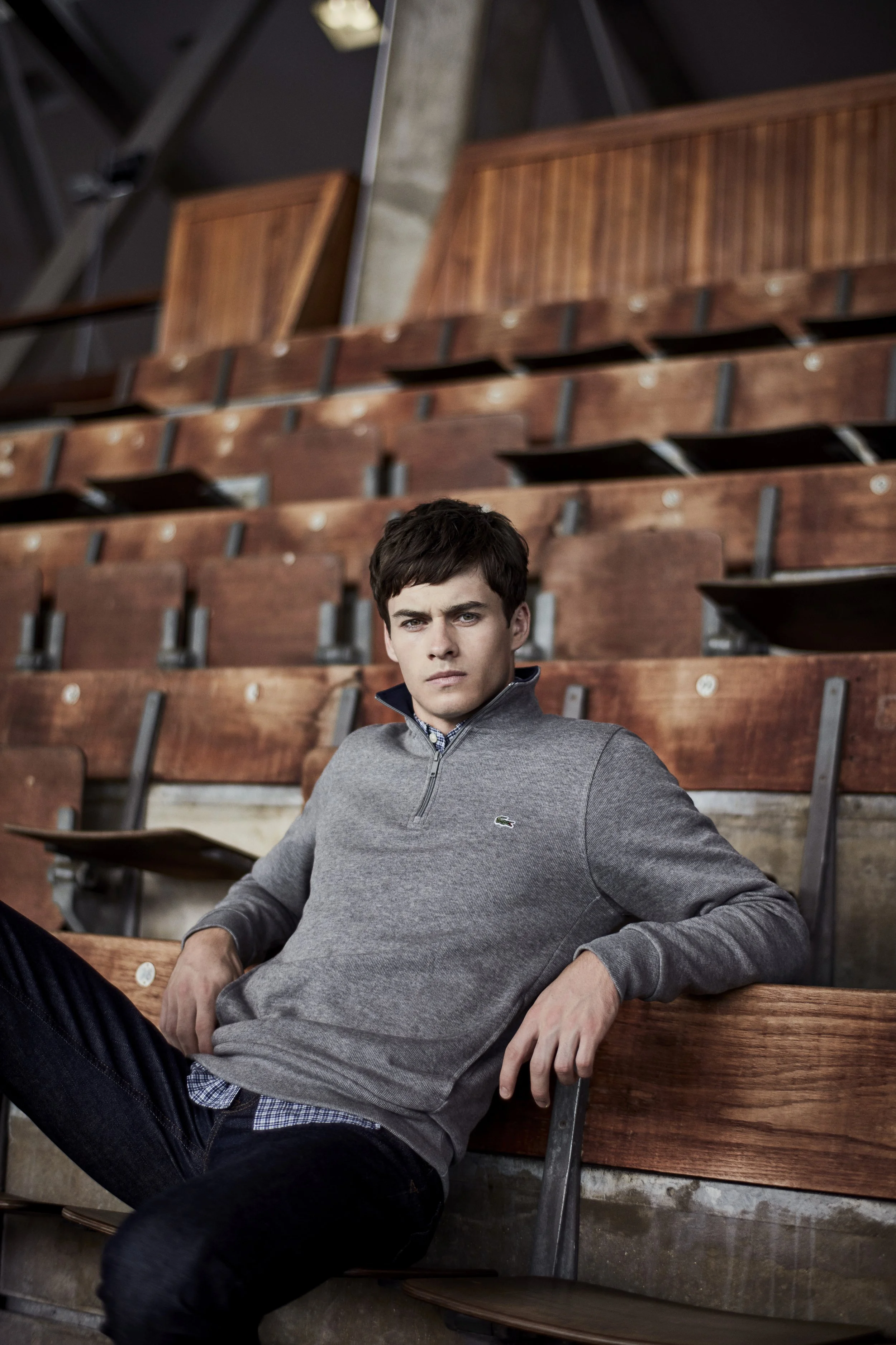 Lacoste, A young man with dark hair, wearing a gray Lacoste sweater over a blue checkered shirt, sitting on wooden bleachers in an indoor gym or auditorium, looking at the camera with a serious expression.