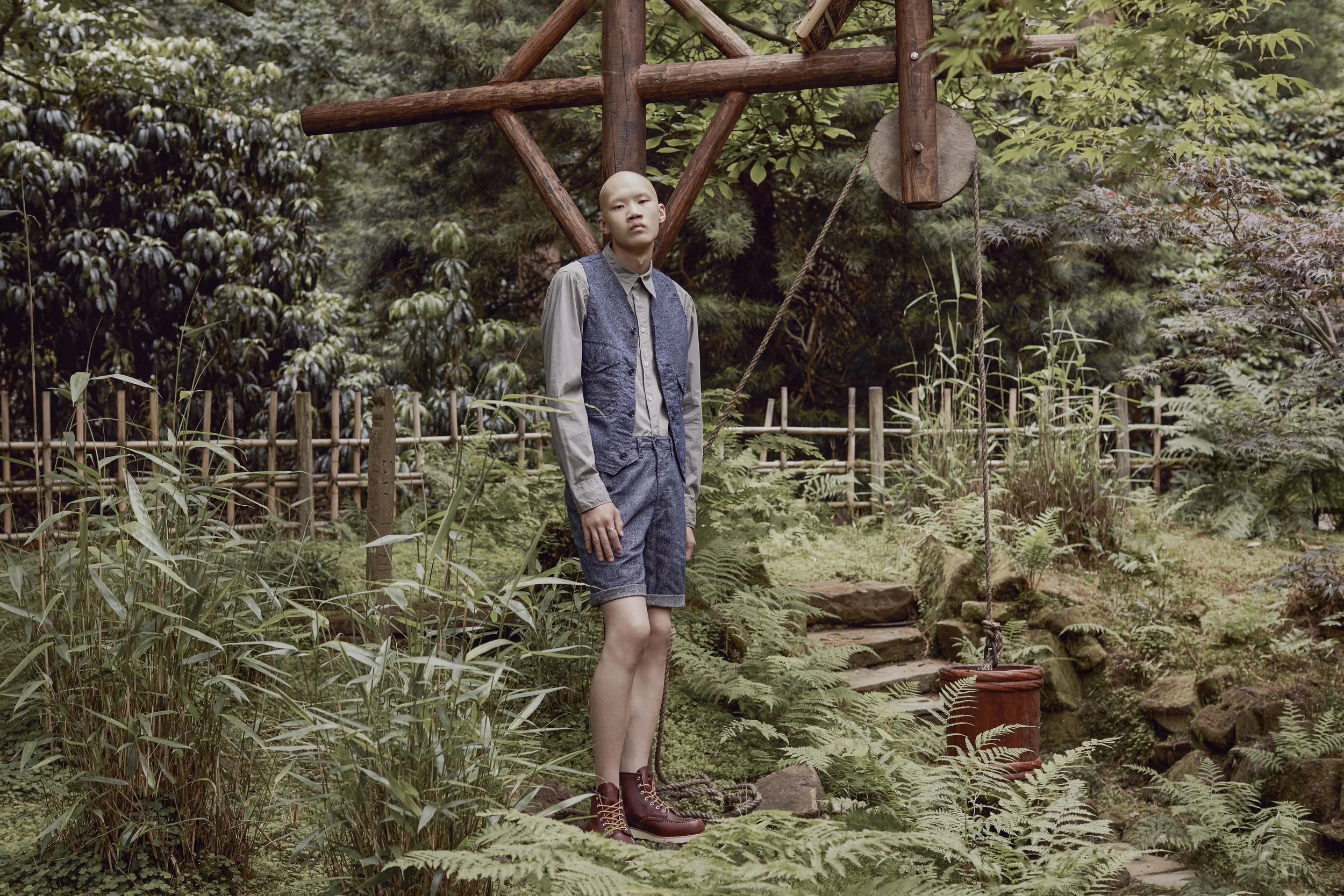 A person with short hair, wearing a denim vest, gray shirt, and rolled-up shorts, standing outdoors on a stone pathway next to a wooden well with a rope and pulley, surrounded by lush greenery and trees.