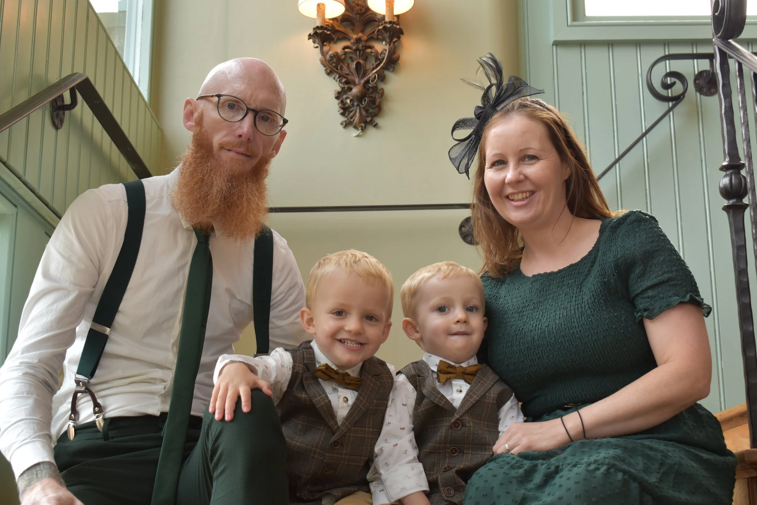 A group of four people, including two young boys and two adults, smile at the camera on a wooden staircase inside a building with green walls and decorative lighting.
