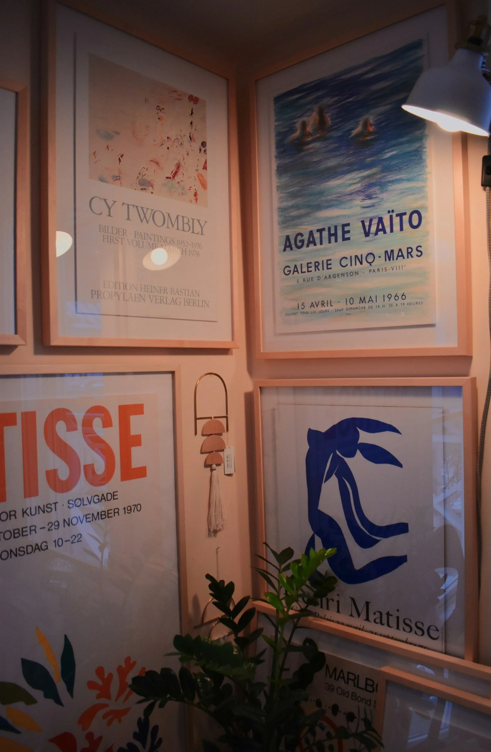 Gallery wall with framed posters advertising art exhibitions, including one for Agathe Vaïto at Galerie Cinq-Mars. There is a small plant in the foreground and a wall lamp illuminating the posters.